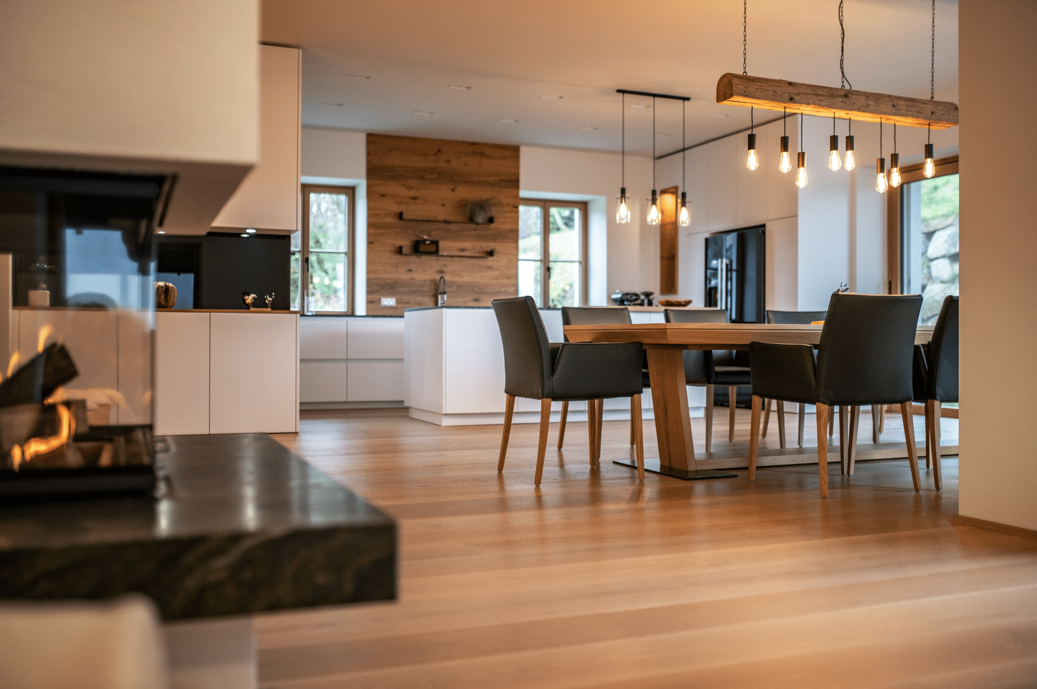 reginaplaza zeigt ein offenes Esszimmer mit Holzboden, großem Holztisch und schwarzen Lederstühlen und angrenzender Küche mit weisser Kochinsel und einem marmoriertem Kamin im Vordergrund.