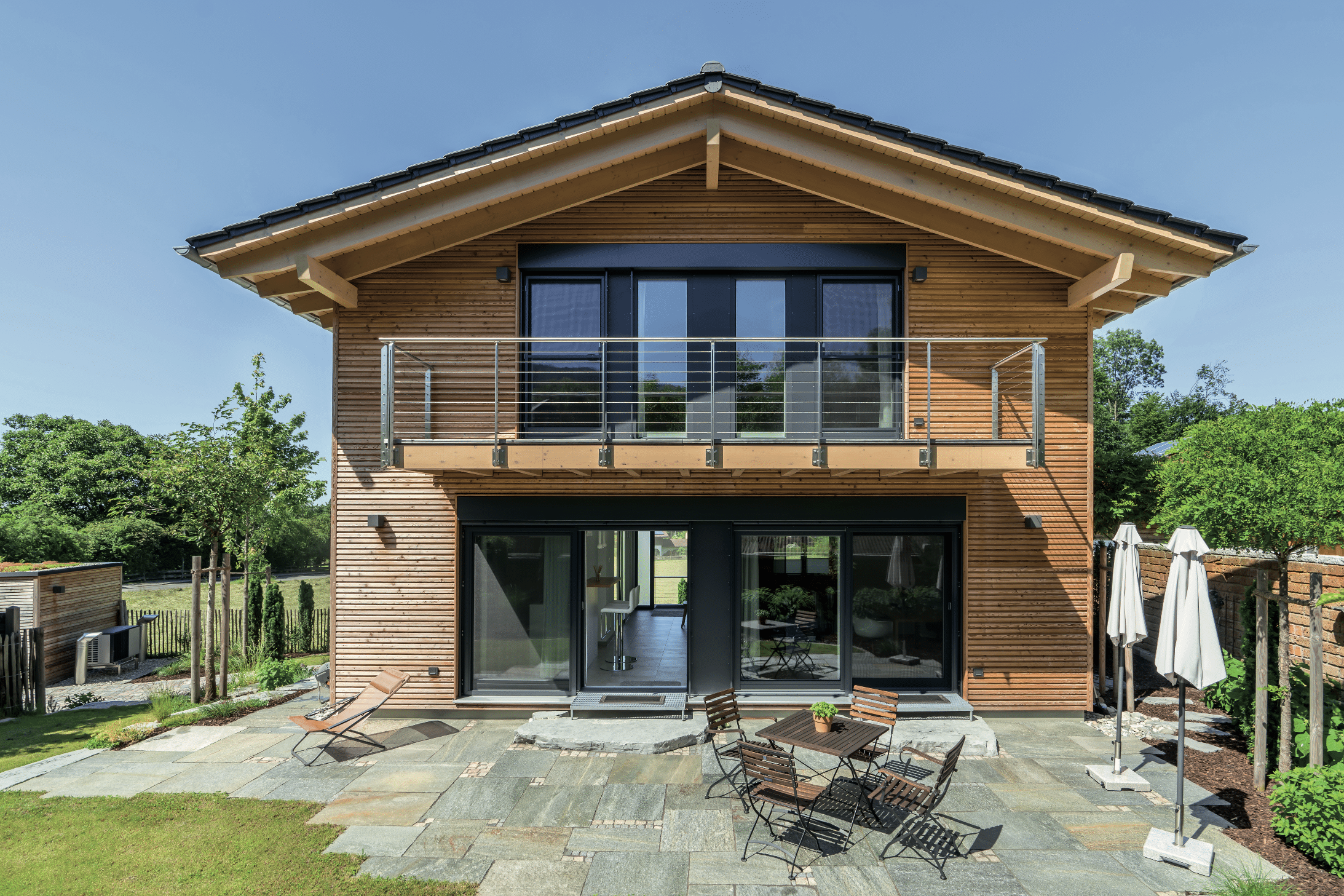 Regnauer zeigt ein Fertighaus aus Holz mit einer Terrasse und großem Garten, Satteldach, Buchsbäumen und einer kleinen Holzhütte.