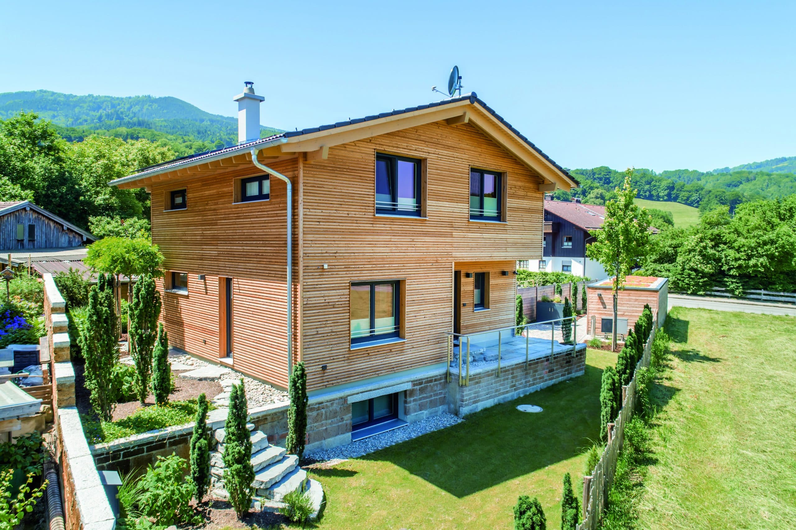 Regnauer zeigt ein Fertighaus aus Holz mit einer Terrasse und großem Garten, Satteldach, Buchsbäumen und einer kleinen Holzhütte.