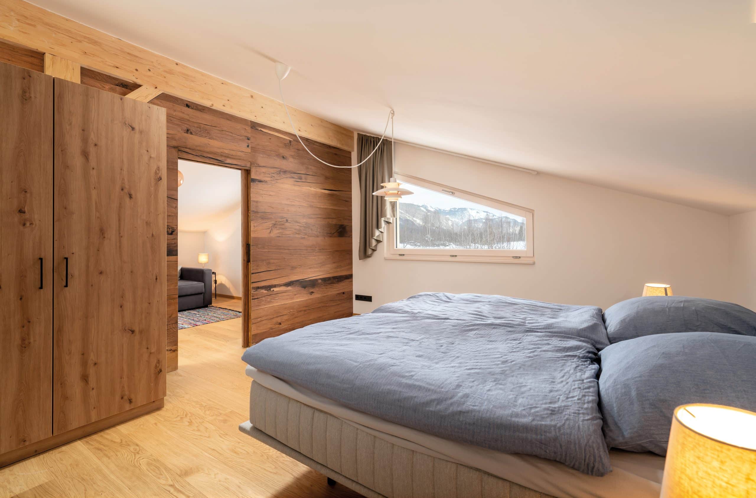 Regnauer zeigt ein Schlafzimmer aus Holz mit Kleiderschrank, Vorhang, und einem Doppelbett mit grauem Überzug.