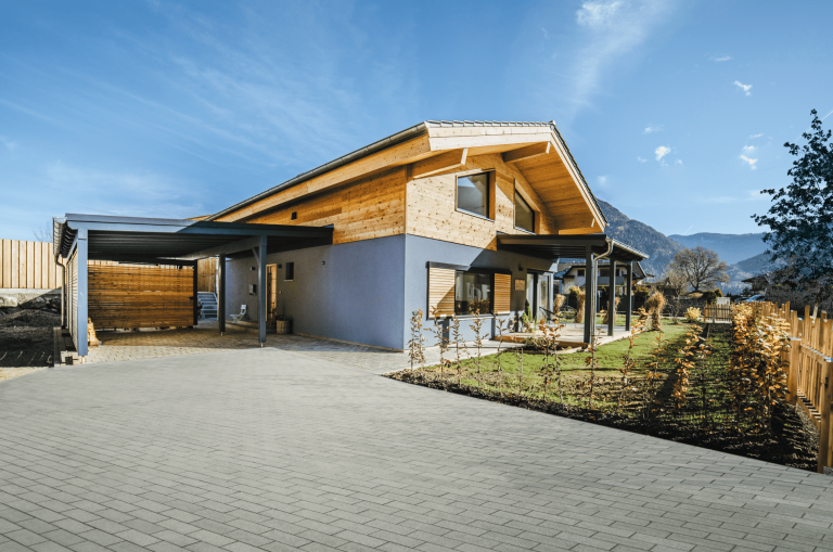 Regnauer zeigt ein einstöckiges Haus mit grauer Fassade und Holzverbau, einem Carport und einem Garten mit herbstlich blühenden Pflanzen.