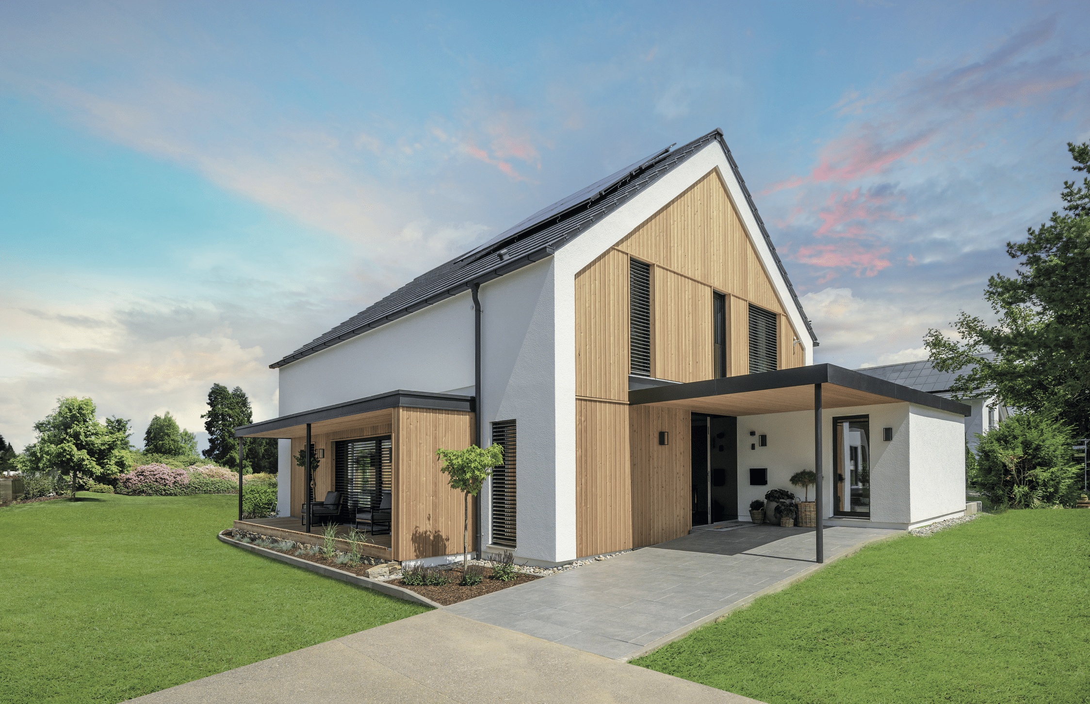 Regnauer zeigt ein modernes, weisses Einfamilienhaus mit heller Holzfassade, überdachter Terrasse, Carport, großer Grünfläche und schwarzem Dach.