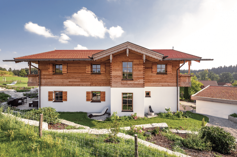 Regnauer Fertighaus zeigt rustikales Einfamilienhaus mit Satteldach mit Holzverziehrung, Holzverkleidung im oberen Stock, überdachtem Balkon und schöner Gartenanlage.