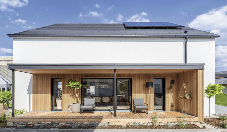 Regnauer Fertighaus zeigt weißes Einfamilienhaus mit Satteldach und großer überdachter Terrasse mit Holzverkleidung.