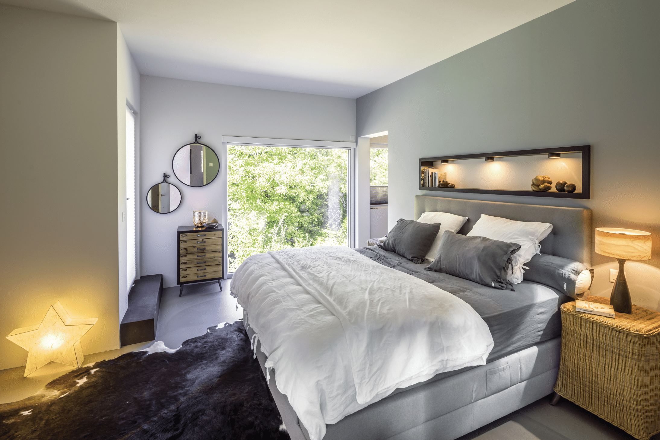 Regnauer zeigt ein Schlafzimmer mit einem Boxspringbett in grau, vielen Kissen, einem Beistelltisch aus Rattan und einem großen Fenster mit Blick ins Grüne.