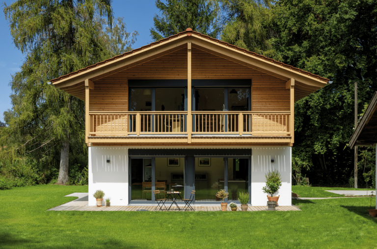 Regnauer zeigt ein Einfamilienhaus im Grünen mit schöner Holzverkleidung, einem Balkon mit Holzgeländer und einer überdachten Terrasse mit Holzboden und Bistro Möbeln.