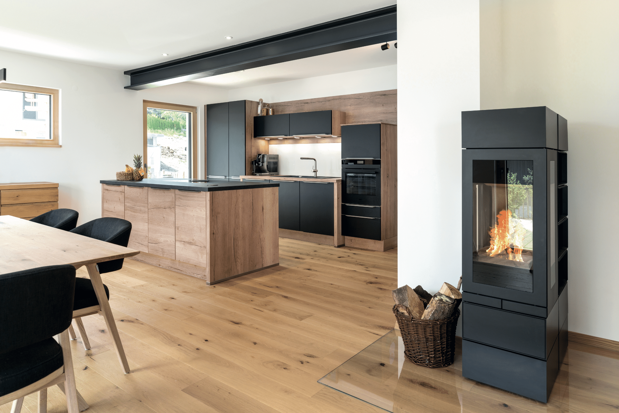Regnauer zeigt eine Küche mit einer Küchenzeile aus Holz und mit schwarzen Fronten, einer Kochinsel mit Kochfeld und dem angrenzenden Esszimmer mit einem schwarzen Kamin in dem ein Feuer für Wärme sorgt.