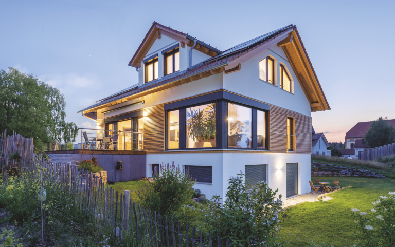 Regnauer zeigt ein weisses Einfamilienhaus mit Holzakzenten, großen Fenstern mit schwarzen Rahmen, einer Terrasse mit einer Treppe die zu einem gepflegten Garten führt.