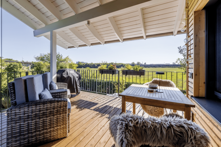 Regnauer zeigt eine Terrasse mit Holzboden, Terrassenüberdachung zum Schutz vor Sonne, Outdoormöbeln aus Rattan und Blick ins Grüne.