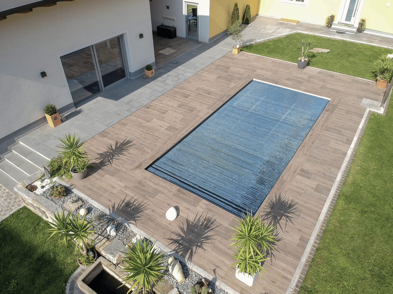Friedrich Reitinger Schwimmbad und Wellness zeigt einen gepflegten Garten mit einem in den Boden eingelassenen, rechteckigen Pool mit einer hellen Holzumrandung und Pflanzen.