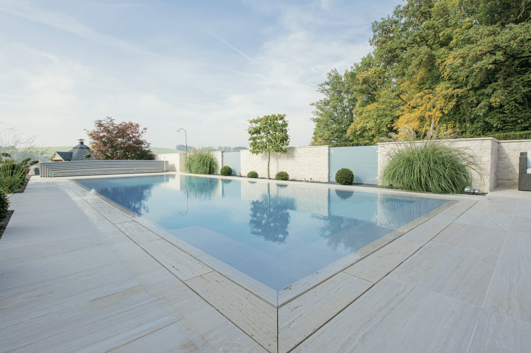 Friedrich Reitinger Schwimmbad und Wellness zeigt einen Garten mit Zaun aus Glas und Ziegelsteinen und einem in den Boden eingelassenen, rechteckigen Pool mit Holzumrandung und Pflanzen.