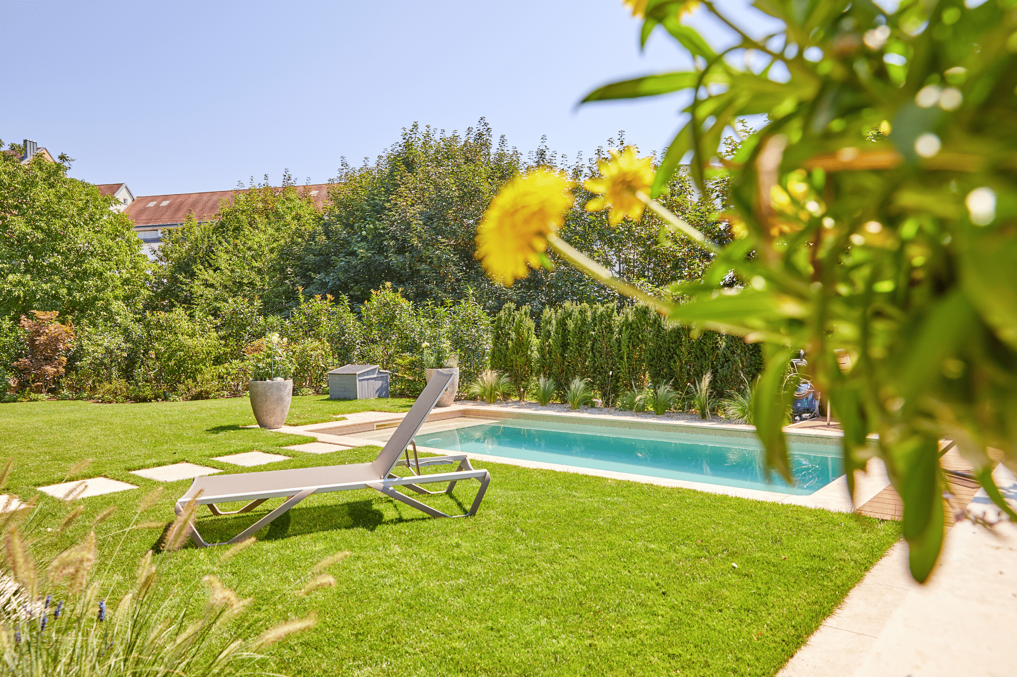 Friedrich Reitinger Schwimmbad und Wellness zeigt einen gepflegten Garten mit einem in den Boden eingelassenen, rechteckigen Pool mit einer gefliesten Poolumrandung und einer Sonnenliege.
