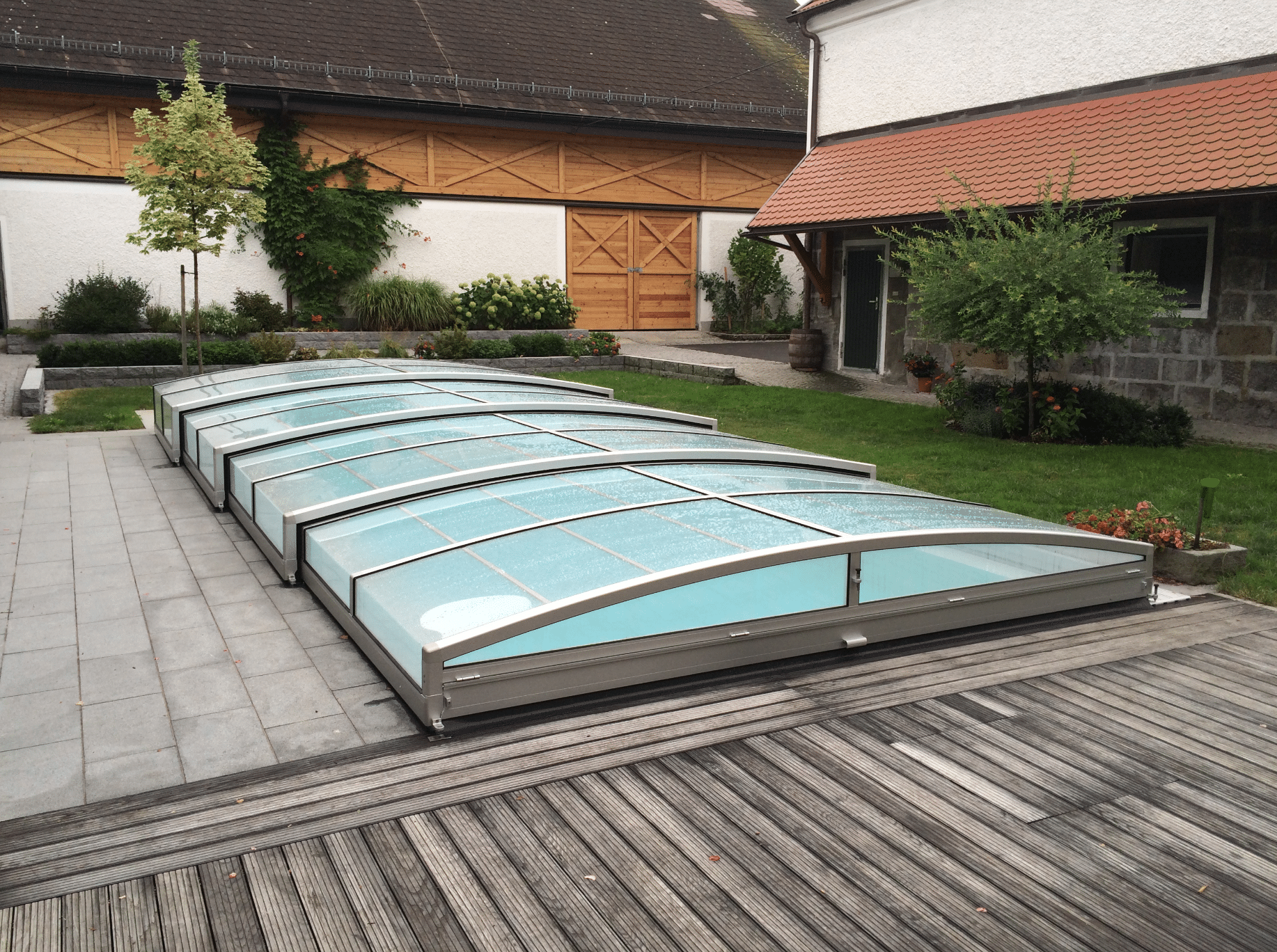 Friedrich Reitinger Schwimmbad und Wellness zeigt einen in den Boden eingelassenen, rechteckigen Pool mit einer Poolüberdachung aus Glas und einer Umrandung aus Holz und Steinplatten.