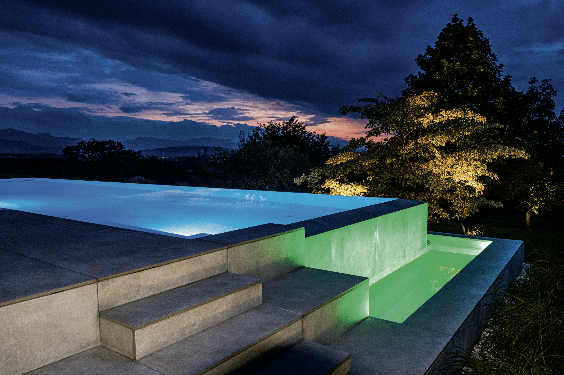 SAUNA-SCHWIMMBAD RUHA Stelzmüller zeigt einen Swimmingpool mit Überlauf bei Nacht mit farbenfroher Beleuchtung.