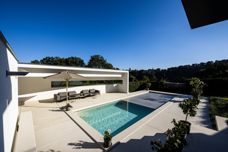 Sonnige Terrasse mit einem großen Swimmingpool von SAUNA-SCHWIMMBAD RUHA Stelzmüller und praktischer Lamellen-Abdeckung.