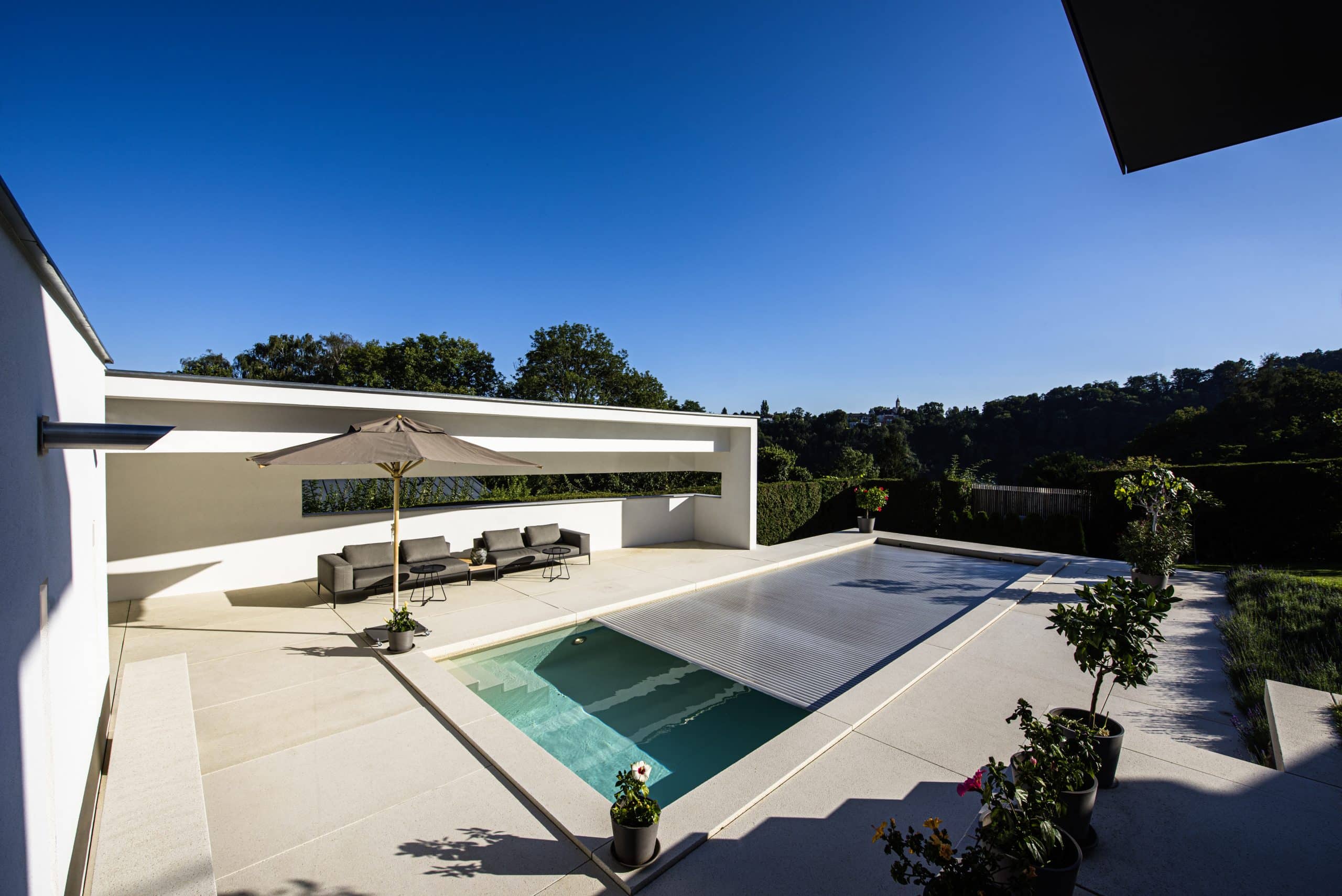 RUHA präsentiert eine Terrasse mit großen beigen Fliesen, einem Pool, einer Lounge und Bepflanzung.