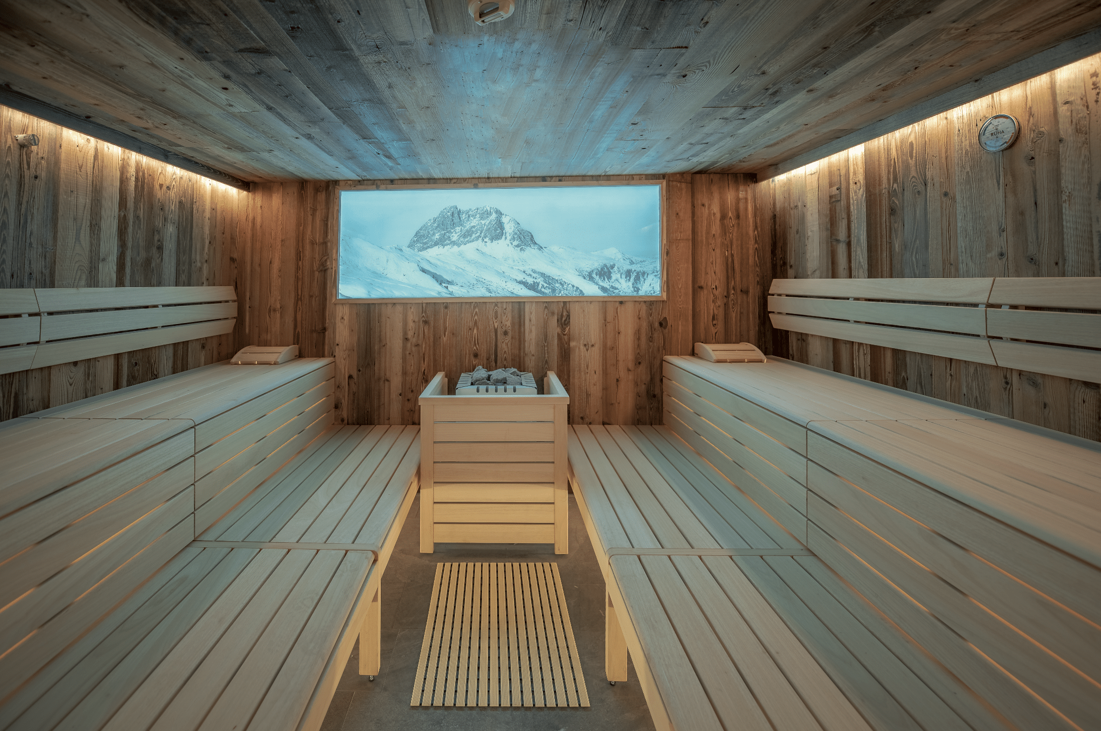 RUHA präsentiert eine Sauna aus Holz von innen, mit dem Aufgussblock vor einem Fenster mit Blick in die Berge.