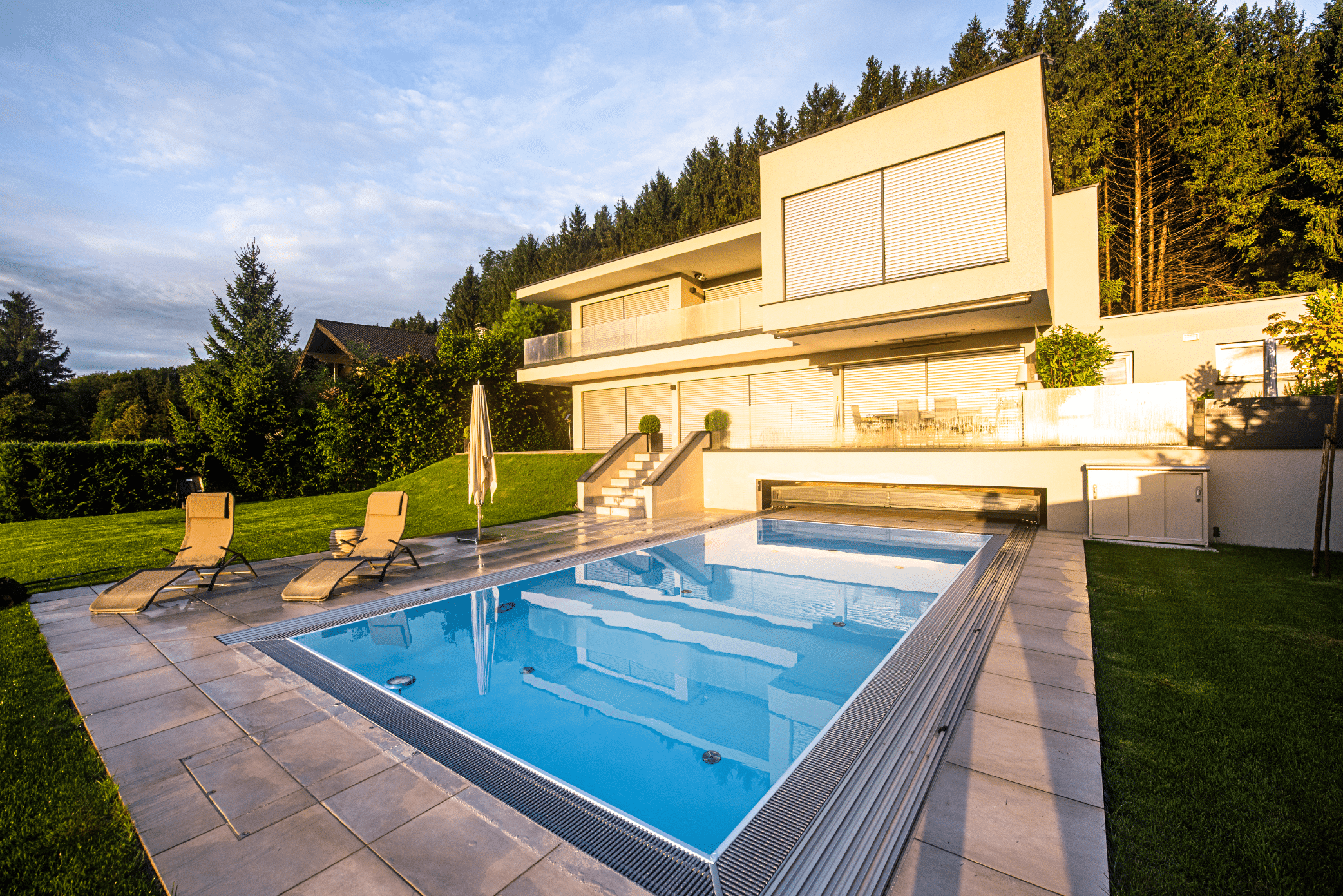 RUHA zeigt einen rechteckigen Pool mit einer Umrandung aus Fliesen und Überdachungsmöglichkeit bei einem Einfamilienhaus mit großem Garten und Sonnenliegen.