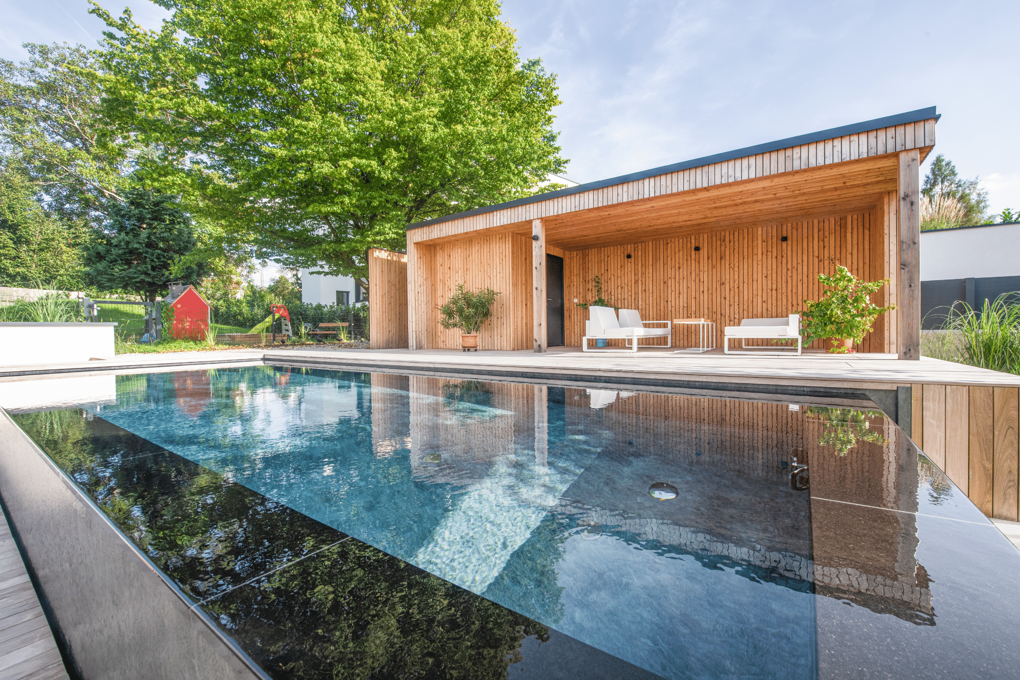 RUHA zeigt einen modernen Infinity Pool mit schwarzem Rand und Holzumrandung in einem Garten mit einer Terrasse, überdachter Sitzlounge und kleinem Spielplatz für Kinder.