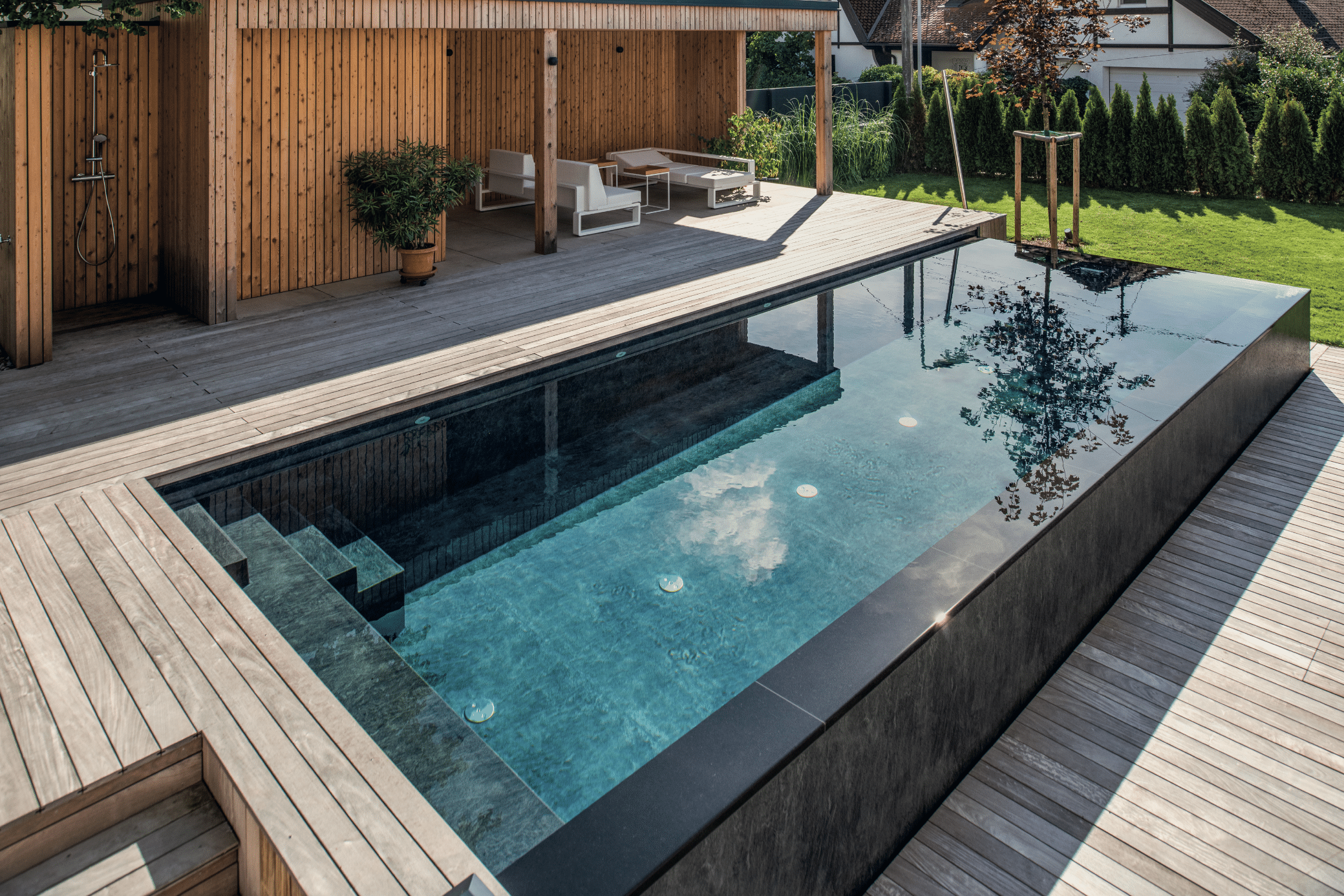RUHA zeigt einen rechteckigen Infinity Pool mit schwarzem Rand und heller Holzumrandung in einem Garten mit einer Terrasse, überdachter Sitzlounge mit gemütlichen Loungemöbeln.