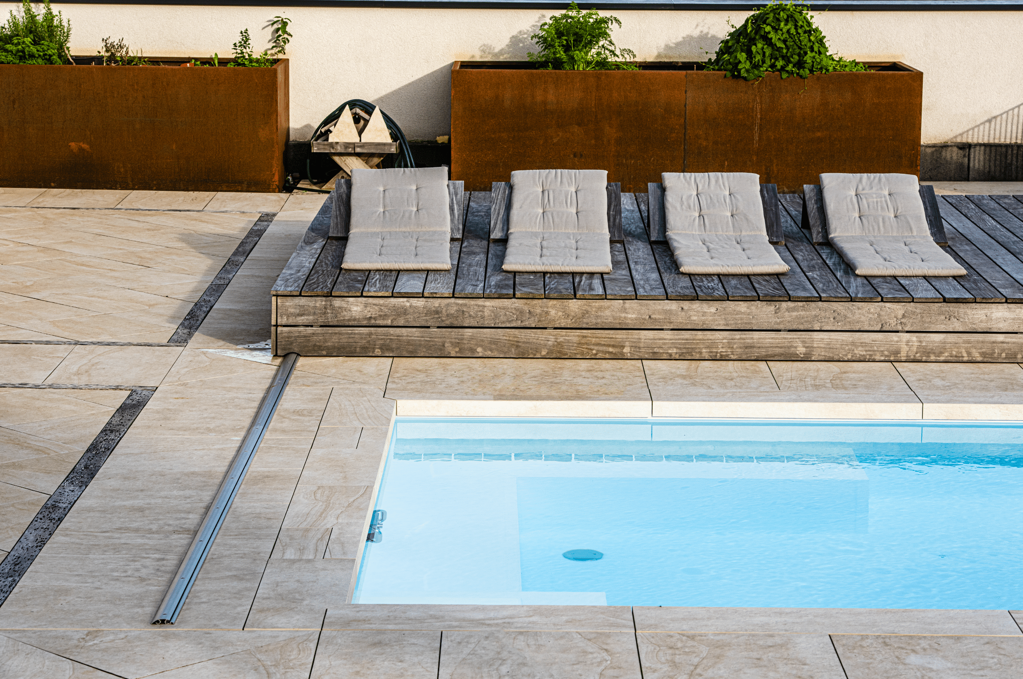 RUHA zeigt einen rechteckigen Pool der im Boden versenkt ist und eine geflieste Poolumrandung hat, neben einem erhöhten Sonnendeck aus Holz mit gemütlichen Polsterauflagen.