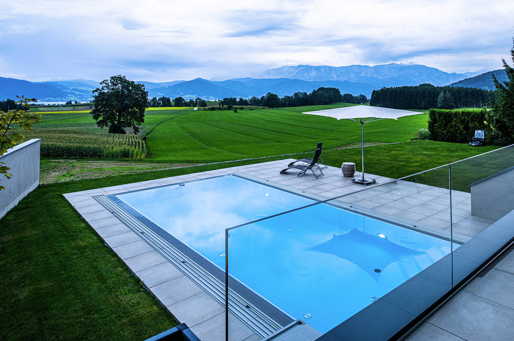 RUHA zeigt einen rechteckigen, im Boden versenkten Pool und der eine geflieste Poolumrandung hat, daneben sind Sonnenliegen und ein Schirm und eine wunderschöne Aussicht ins Grüne.