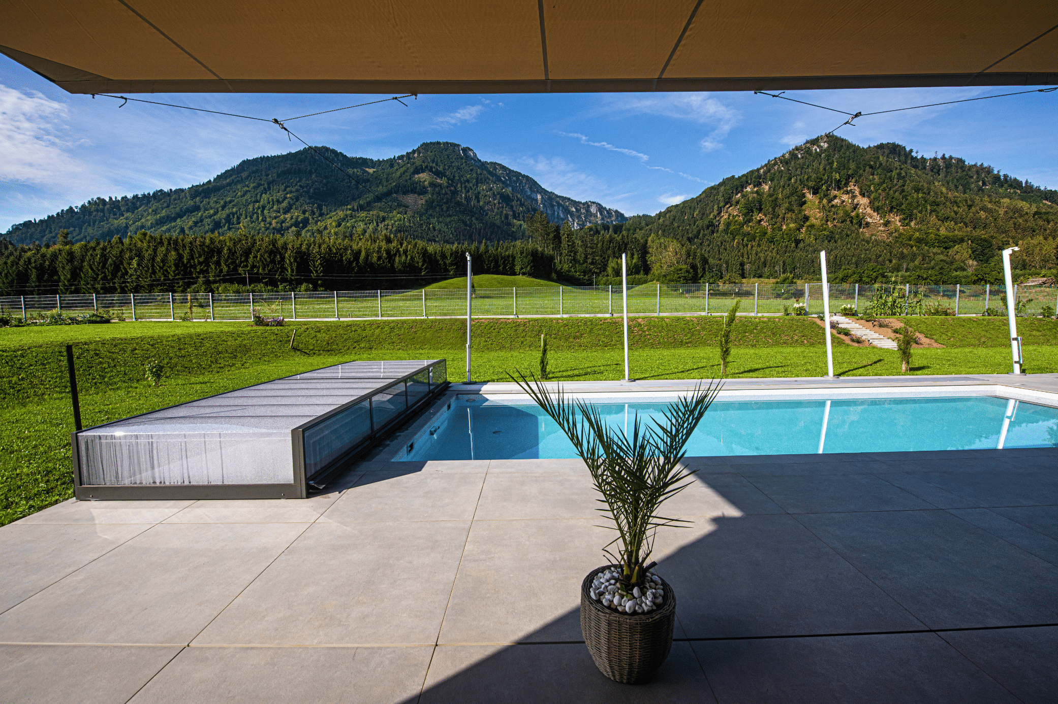 RUHA zeigt einen rechteckigen Pool mit einer Poolüberdachung aus Glas und einer gefliesten Poolumrandung auf einer Terrasse mit Markise.