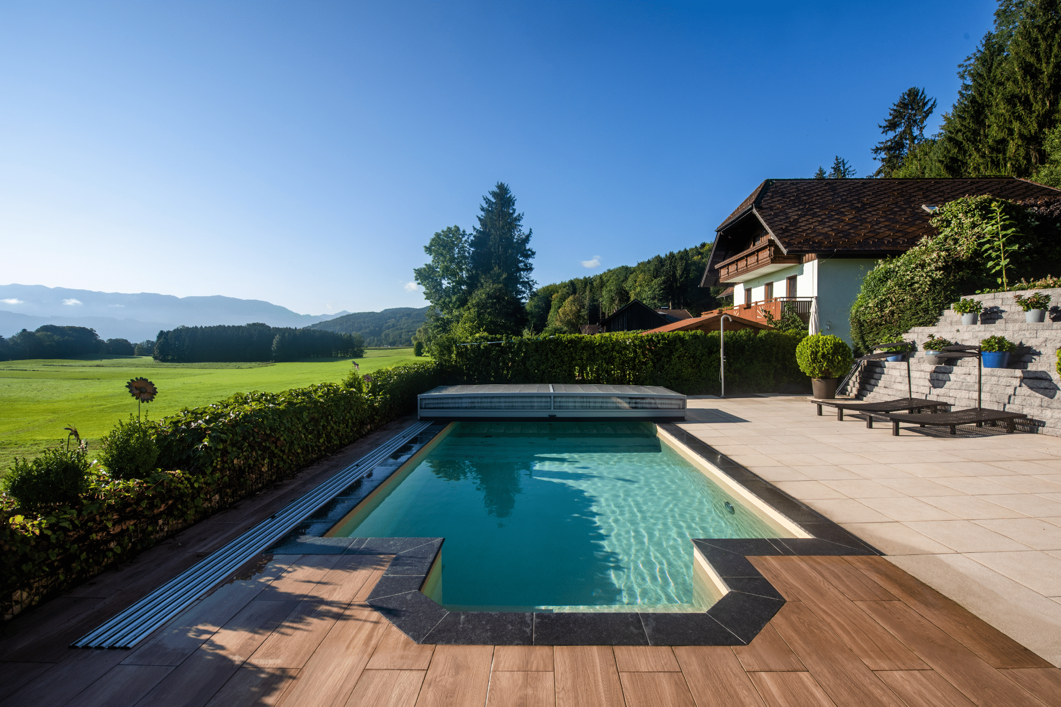 RUHA zeigt einen rechteckigen Pool mit rundem Bereich für den Einstieg, Stufen und einer Poolüberdachung aus Glas zum Schieben auf einer gefliesten Terrasse mit Sonnenliegen.