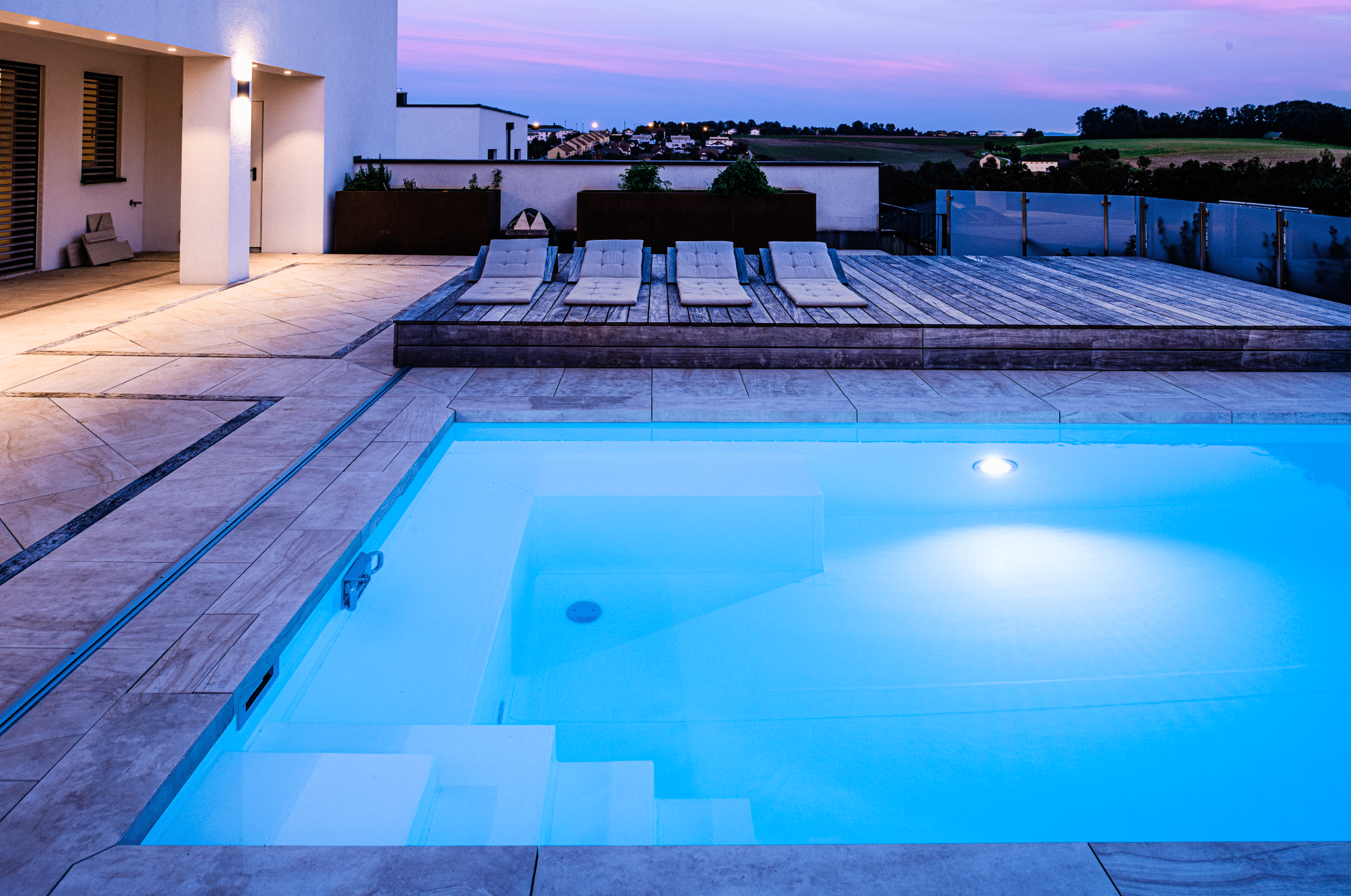 RUHA zeigt einen rechteckigen Pool der im Boden versenkt ist und eine geflieste Poolumrandung hat, neben einem erhöhten Sonnendeck aus Holz mit gemütlichen Polsterauflagen in der Abenddämmerung.