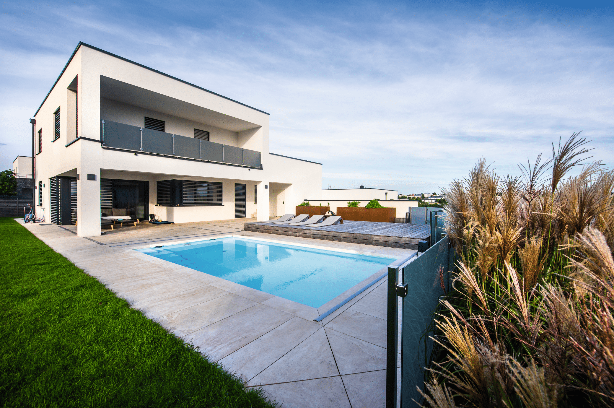 RUHA zeigt einen im Boden versenkten rechteckigen Pool mit gefliester Umrandung und einem Sonnendeck aus Holz mit gemütlichen Liegen in einem Garten eines Einfamilienhauses mit überdachter Terrasse.