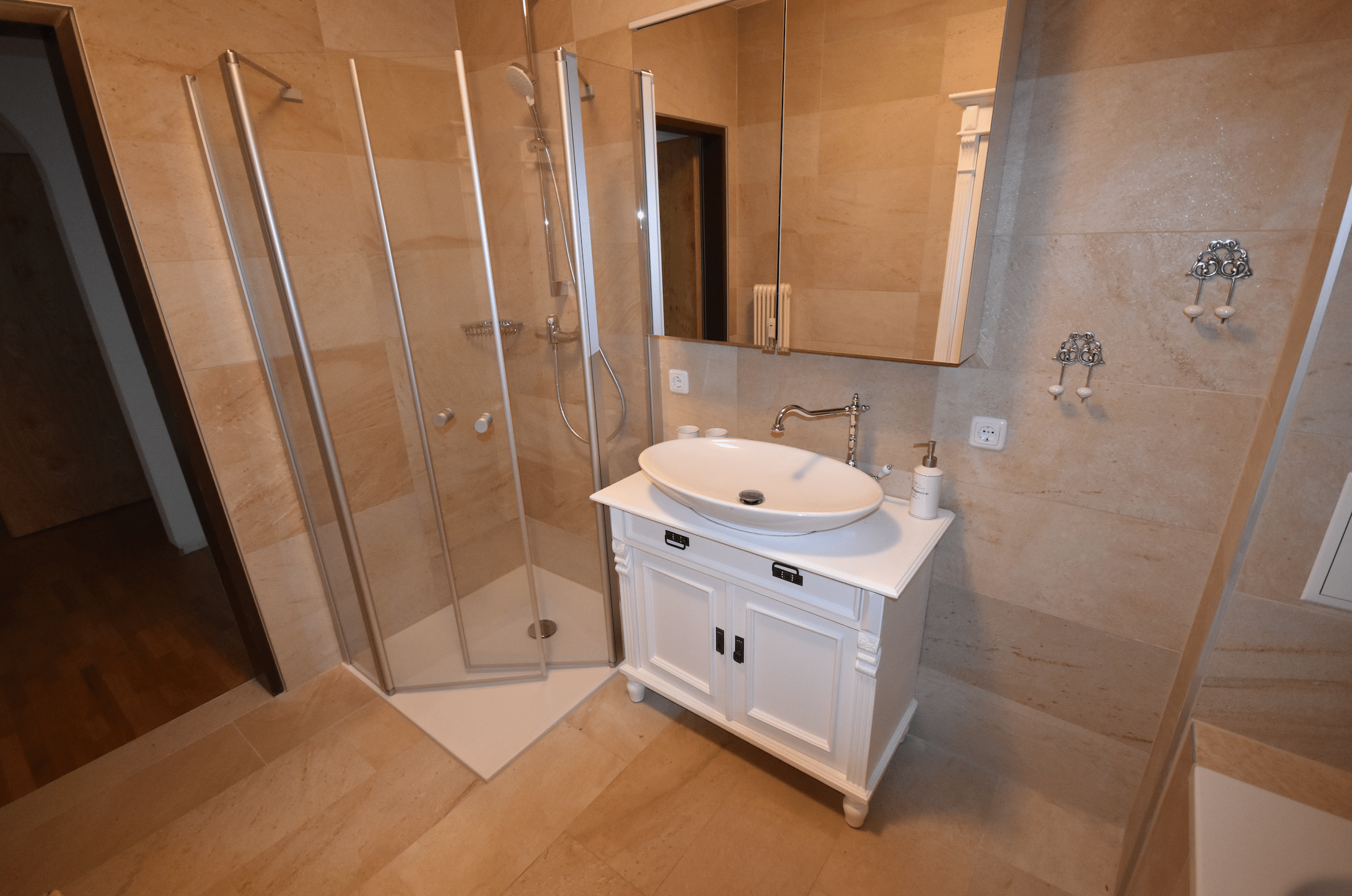 Saniton zeigt ein helles gefliestes Badezimmer mit einer Eckdusche, einem weissen Waschbecken mit Unterschrank und silberner Armatur.