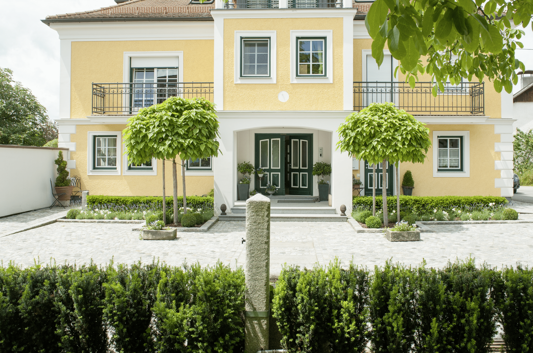 Gartenbau Schmid zeigt die Zufahrt eines großen gelben Hauses mit niedrigen Hecken und zwei Bäumen neben rechts und links neben der Haustüre.
