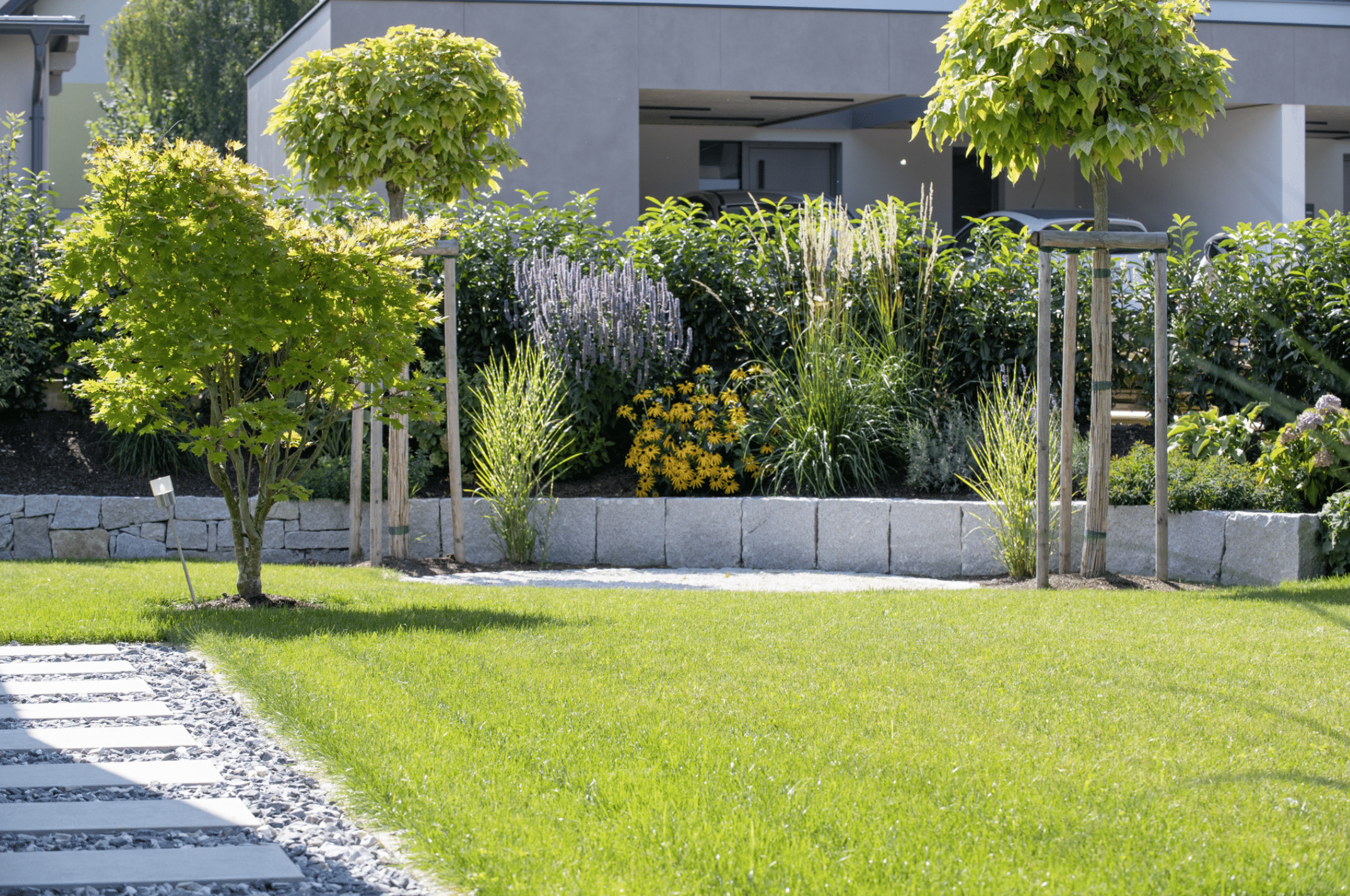 Gartenbau Schmid zeigt ein bepflanztes Beet aus Steinen mit dichter Hecke dahinter als Sichtschutz, angelegten Weg und gepflegtem Rasen.