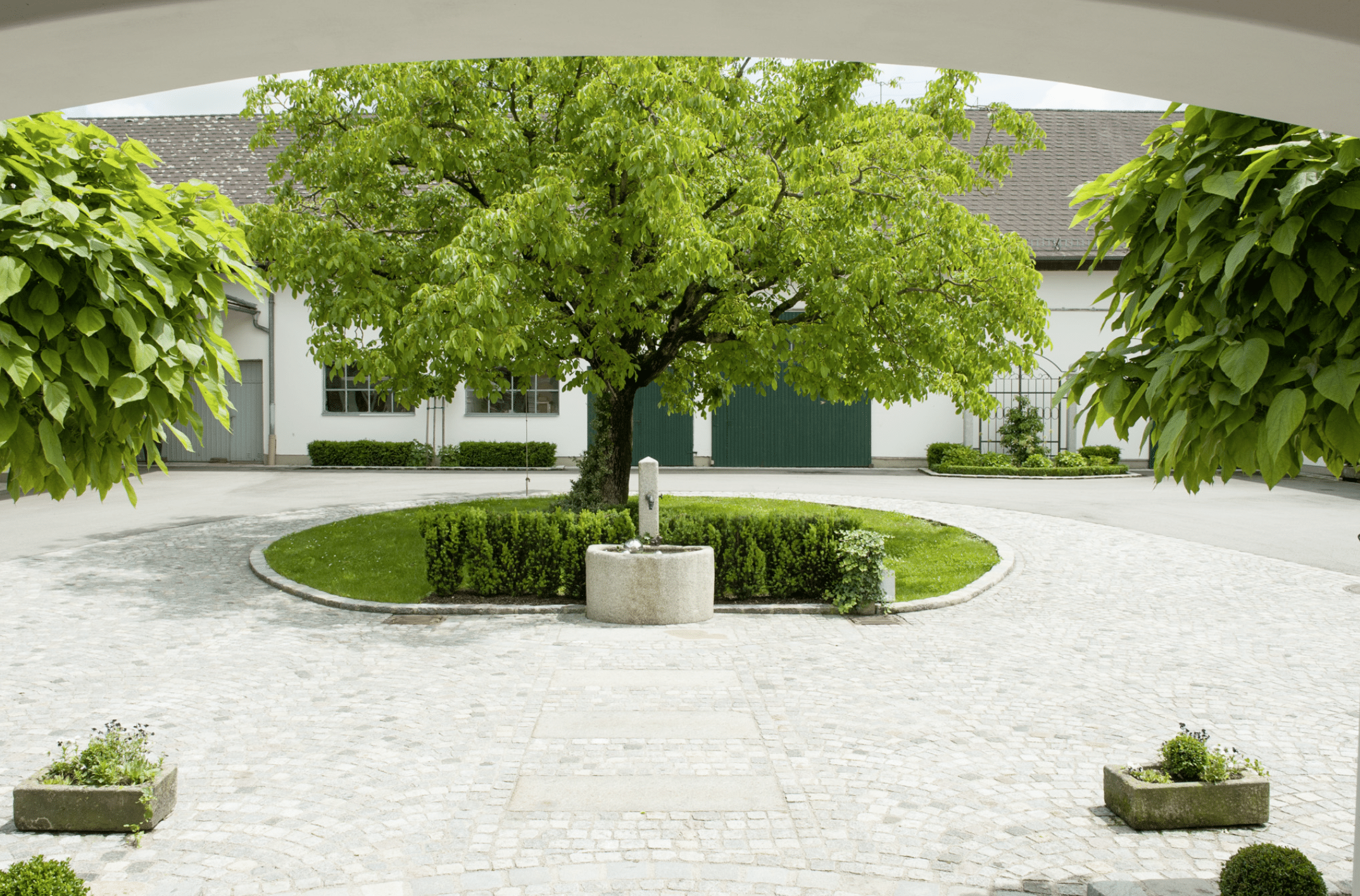 Gartenbau Schmid präsentiert dem gepflasterten Innenhof eines Bauernhofs mit hohem Baum und Brunnen in der Mitte.