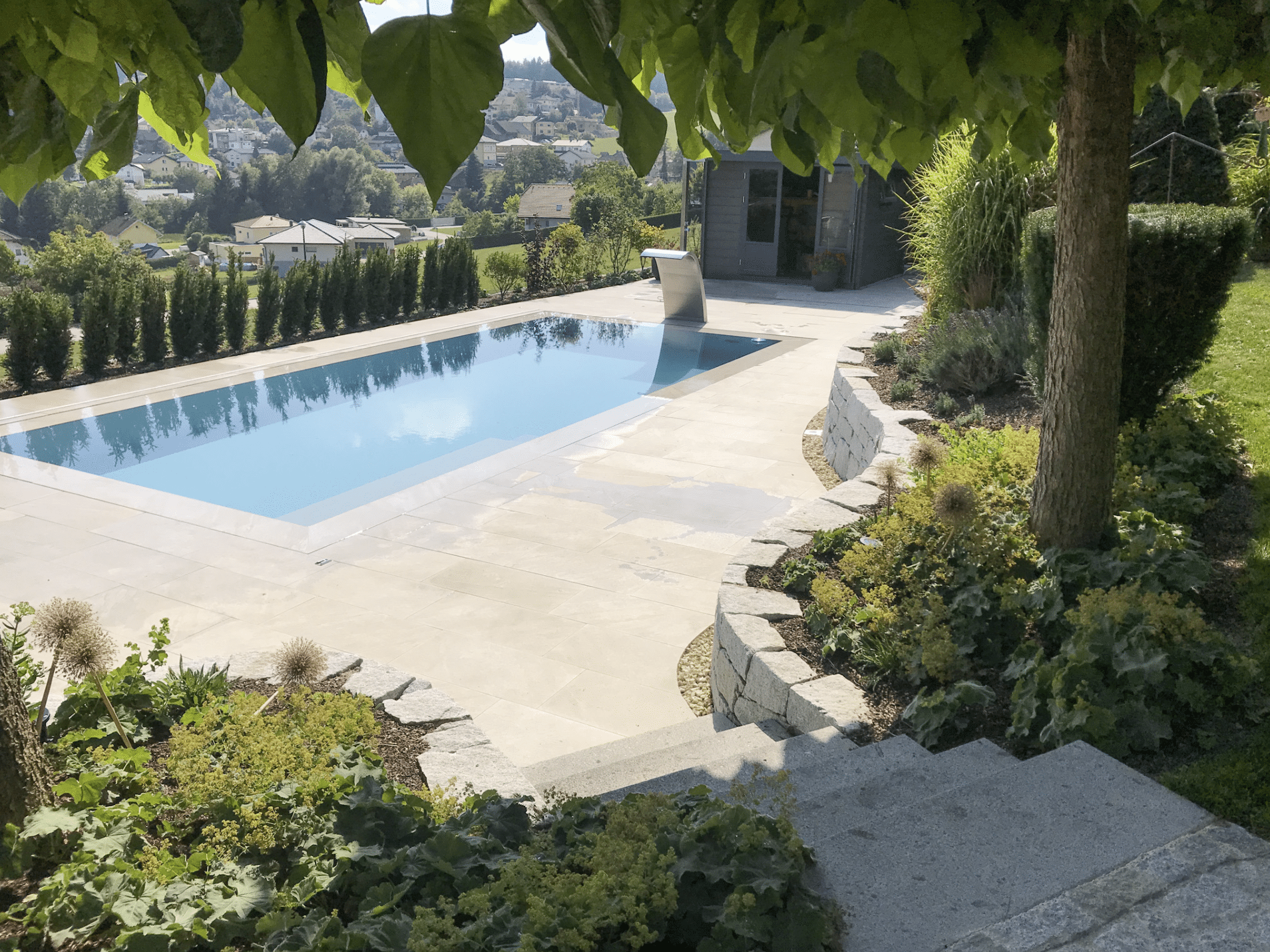 Gartenbau Schmid zeigt eine Terrasse mit Steinfliesen, Pool und einer Treppe in den Garten mit verschiedenster Pflanzen.