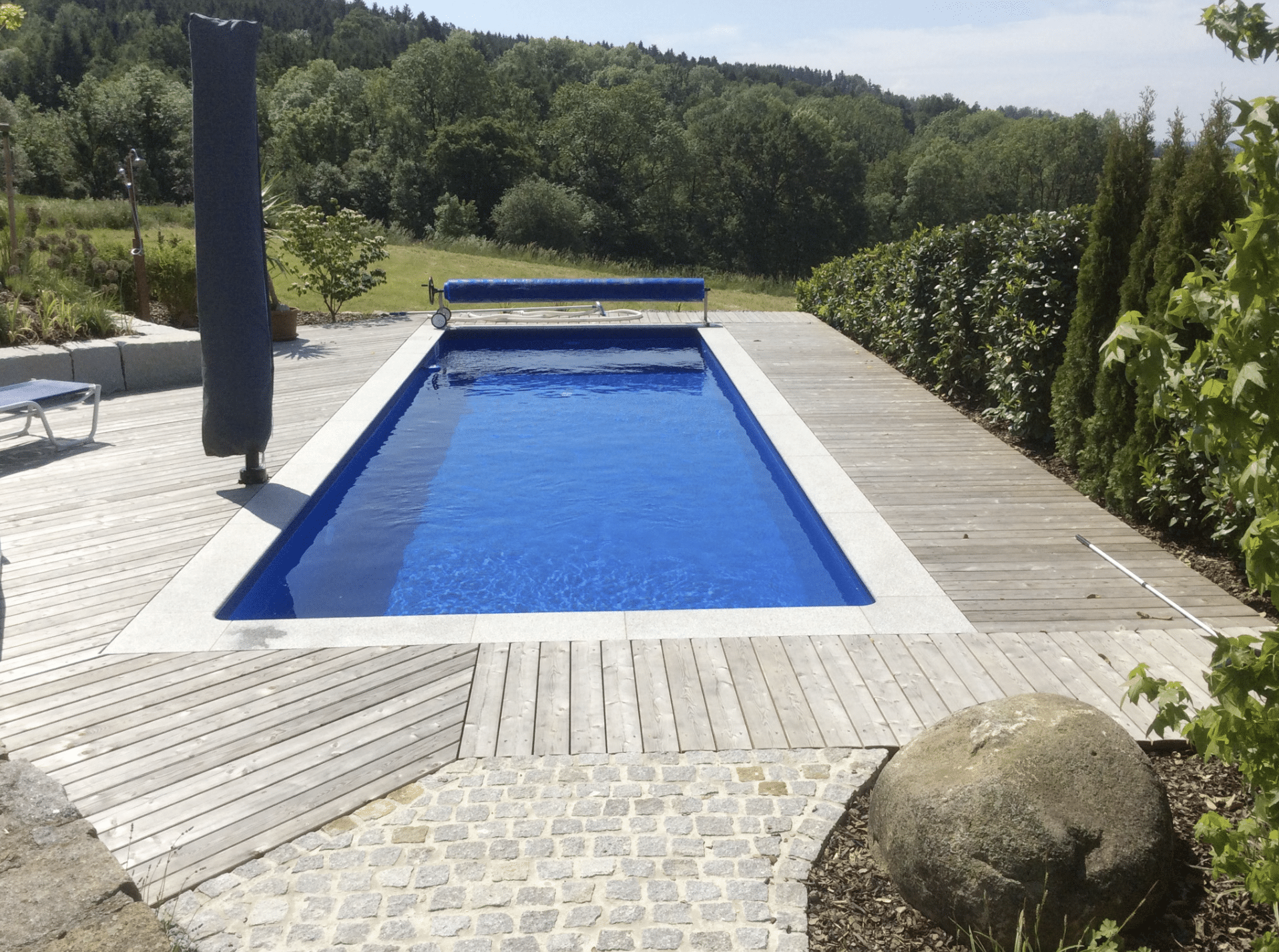 Gartenbau Schmid zeigt einen rechteckigen Pool mit Holzverbau rundeherum und verschiedene Hecken als Sichtschutz.