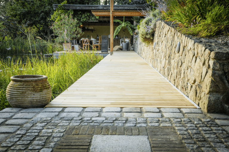 Gartenbau Schmid zeigt eine große Terrasse mit Naturpool und langem Holzweg, Outdoorküche und Steinmauer.