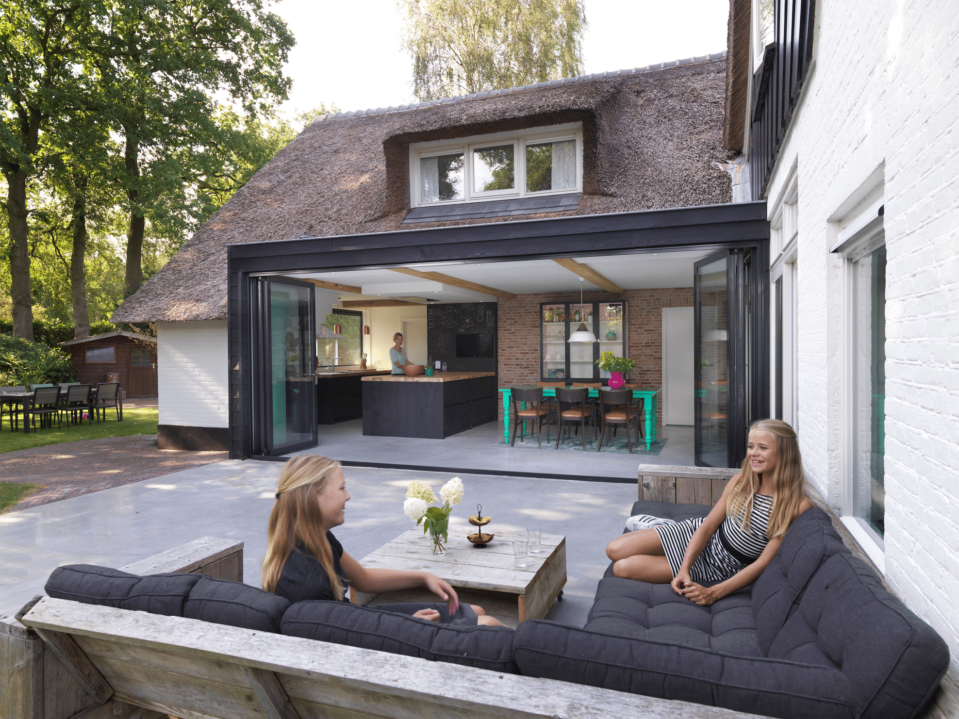 Wohnhaus mit Reetdach und Terrasse mit Küche und Essbereich in einem Zubau in Wintergarten-Optik mit einer Glasfront bestehend aus deckenhohen Falt-Schiebetüren von Solarlux.