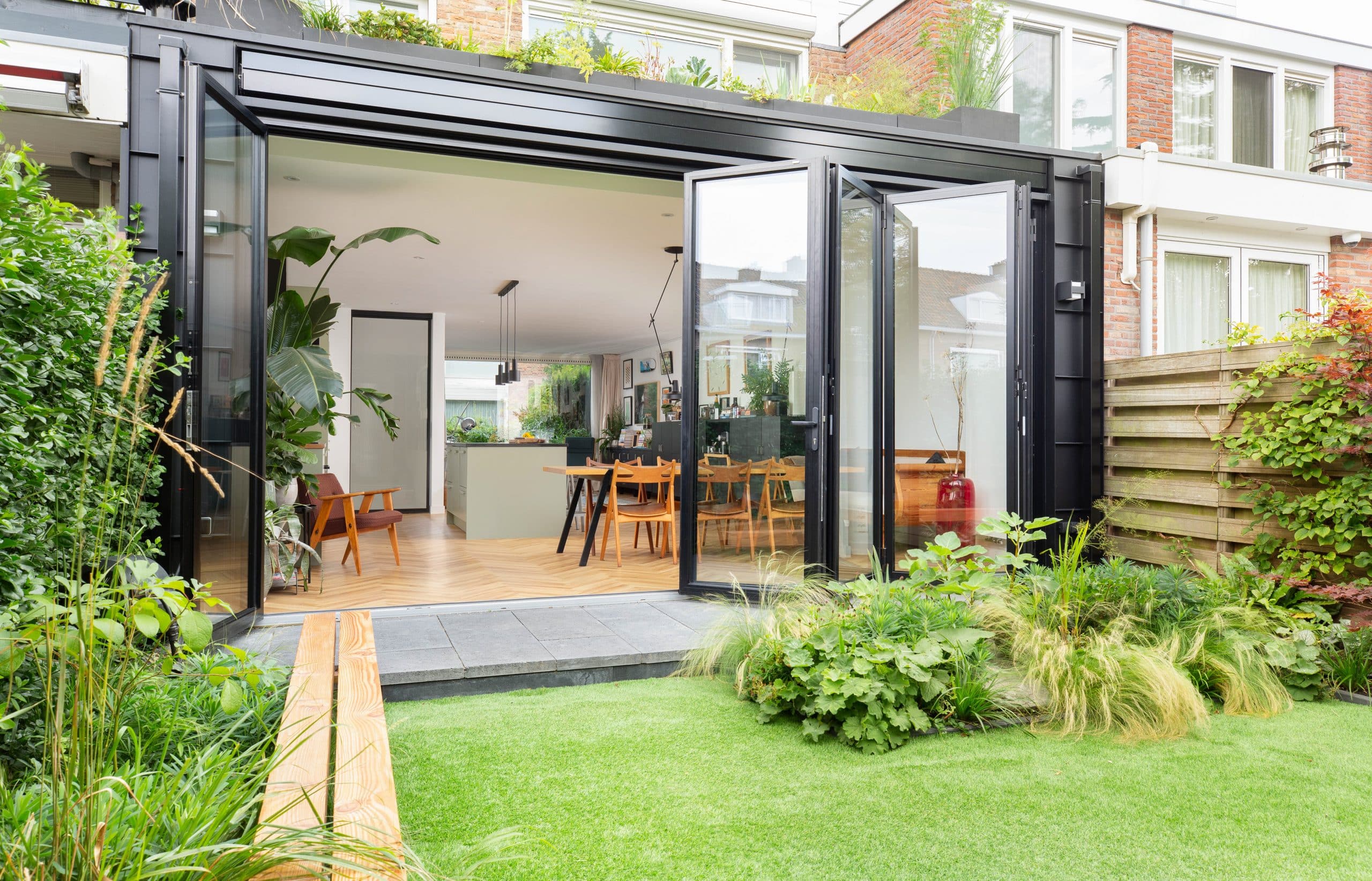 Solarlux zeigt eine Glasfaltwand die Esszimmer und Garten trennt, im Garten sind Pflanzen und eine kleine Terrasse.