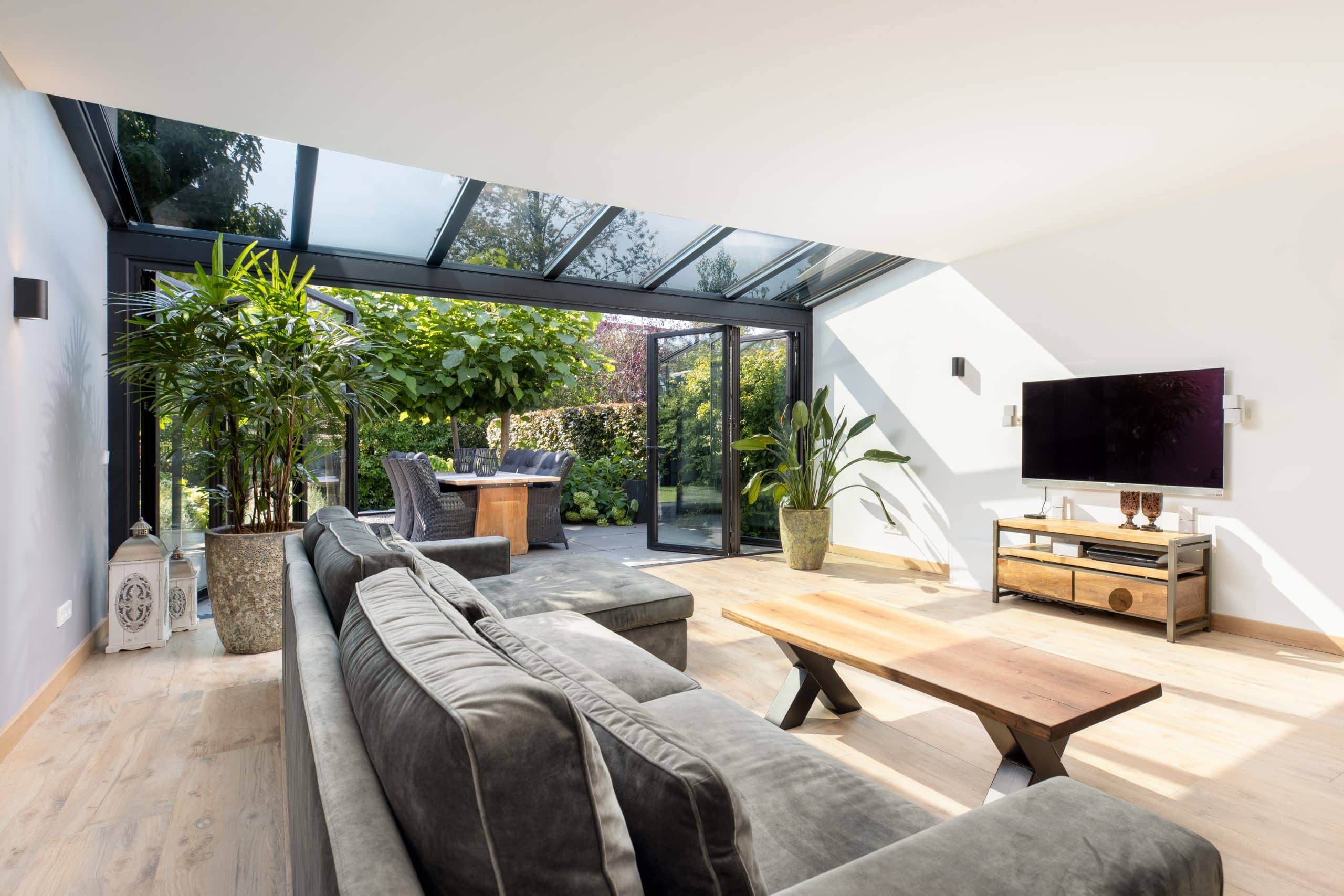 Solarlux zeigt ein Wohnzimmer mit Couch, Fernseher, Tisch und passender Dekoration vor einer Glasfaltwand die in den Garten führt.