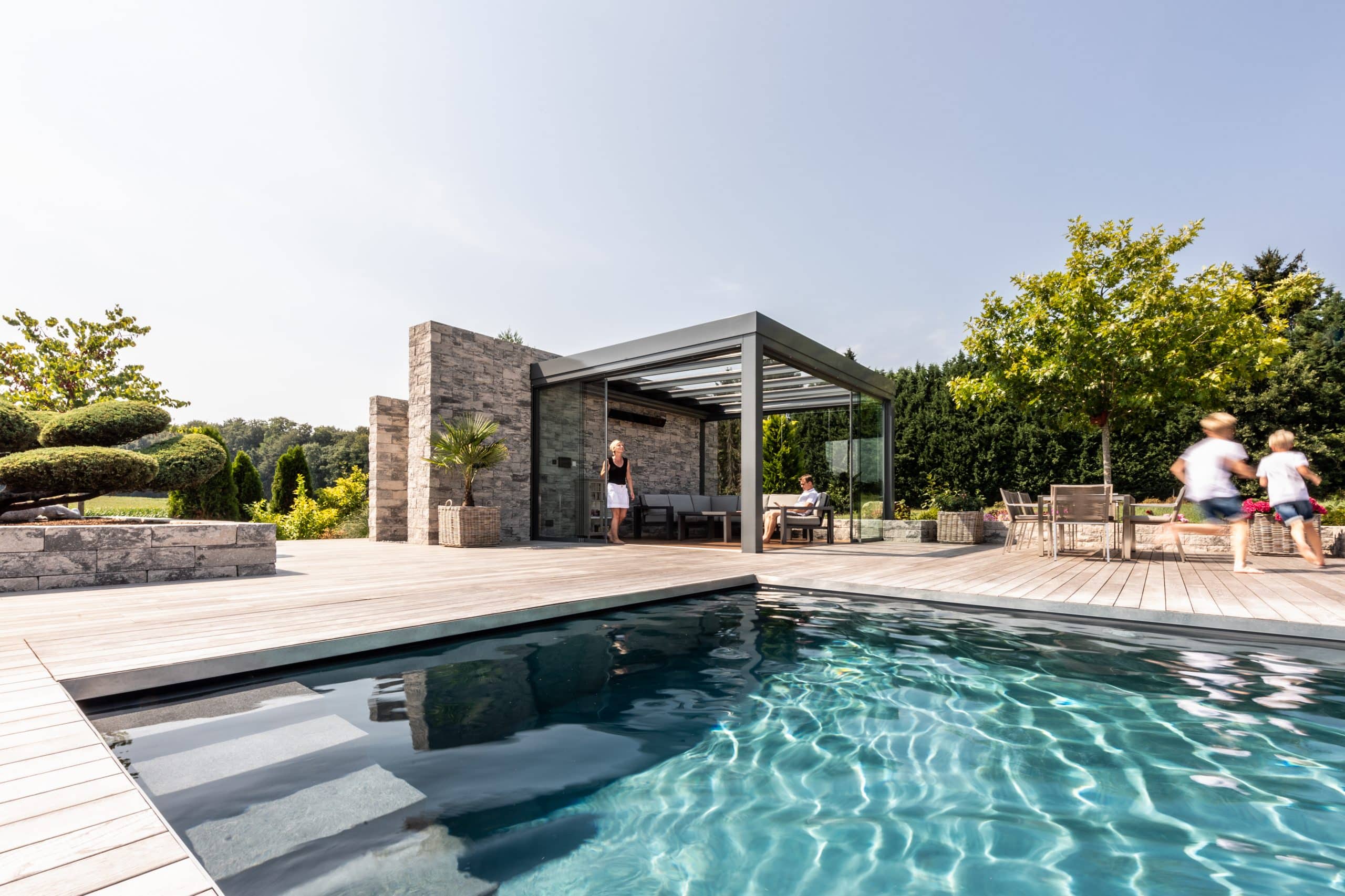 Solarlux zeigt ein barrierefreies Poolhaus aus Glas auf einer Terrasse aus Holz und einigen Steinmauern.
