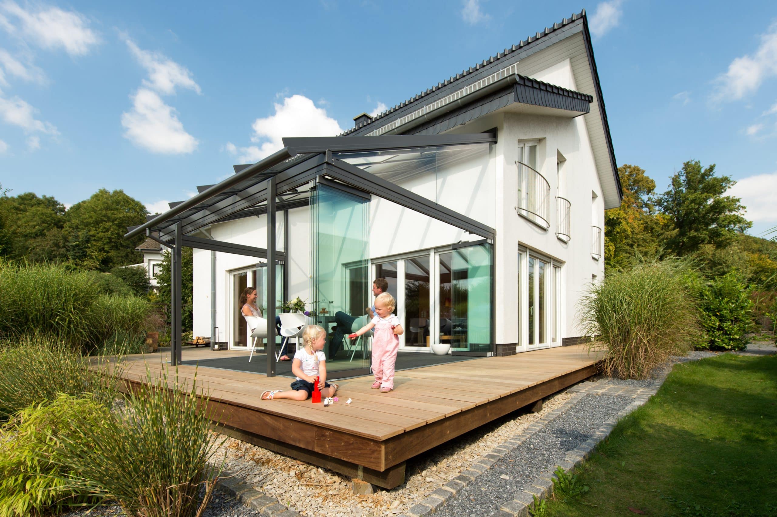 Solarlux zeigt ein modernes,weißes Einfamilienhaus mit großer Glasfaltwand zur Terrasse raus auf der Gartenmöbel stehen und Kinder spielend den Garten sehen.