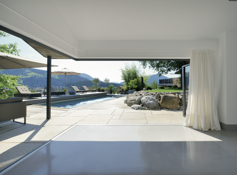 Solarlux zeigt modernen offenen Wohnraum mit Glasfaltwänden mit Blick auf Terrasse mit Pool und Steinmauer.