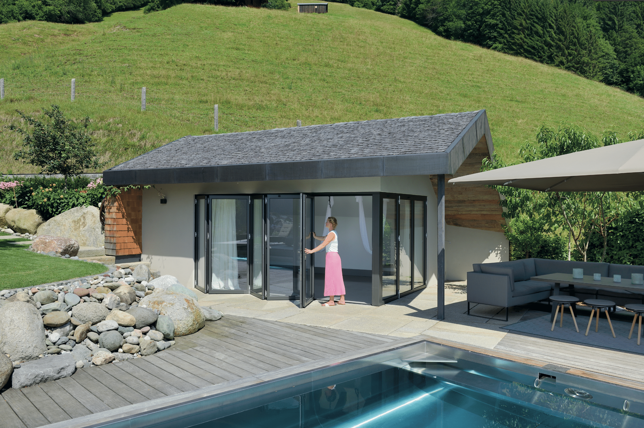 Junge Frau öffnet die moderne Glasfaltwand von Solarlux am modernen Poolhaus und exklusivem Edelstahlbecken mit Terrassenumrandung aus Holz in herrlicher Naturlandschaft.