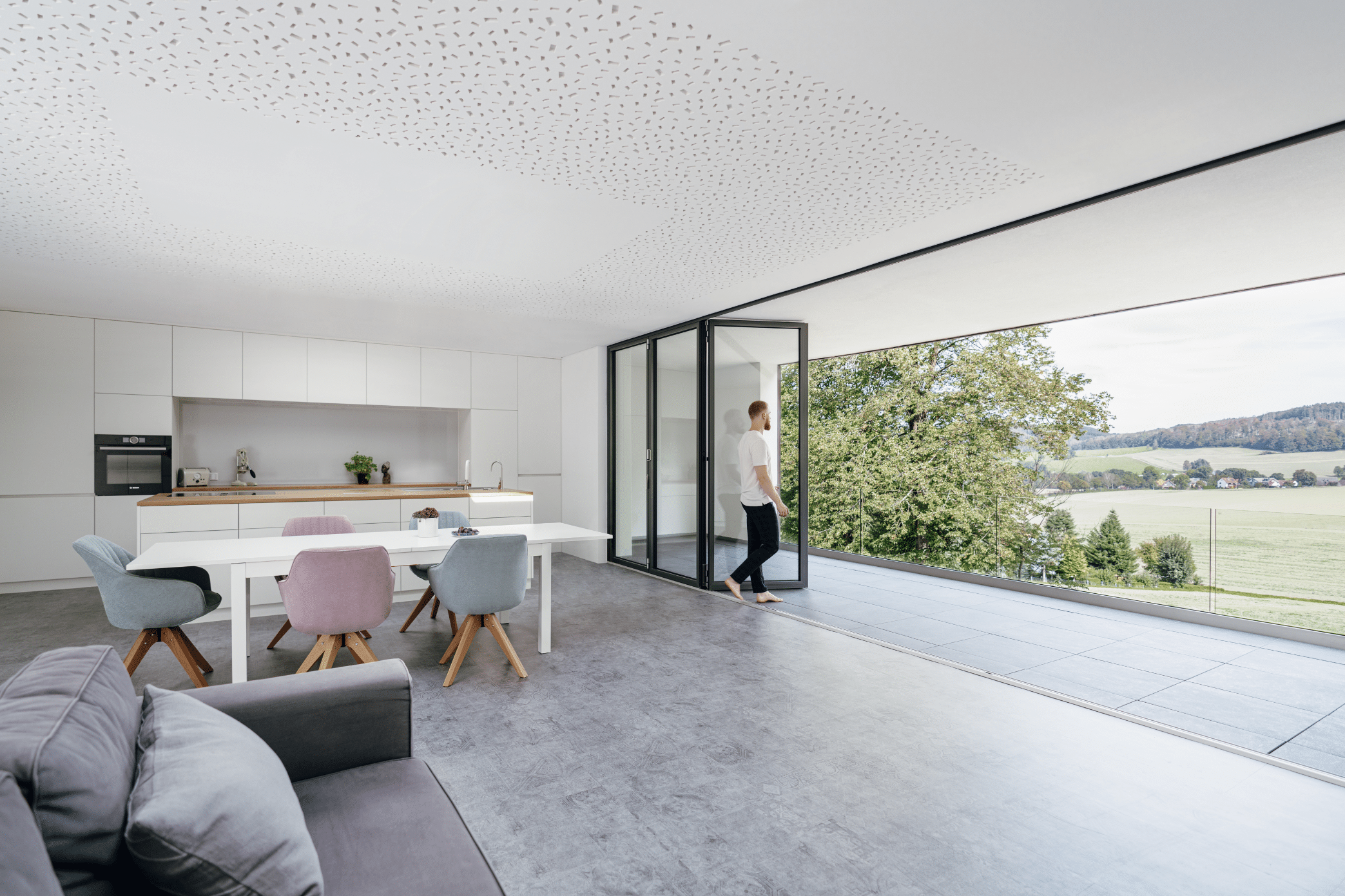 Solarlux zeigt einen offenen Wohnbereich mit Küche, Essbereich und einer grauen Couch mit einer Glasfaltwand und überdachtem Balkon mit Aussicht ins Grüne.