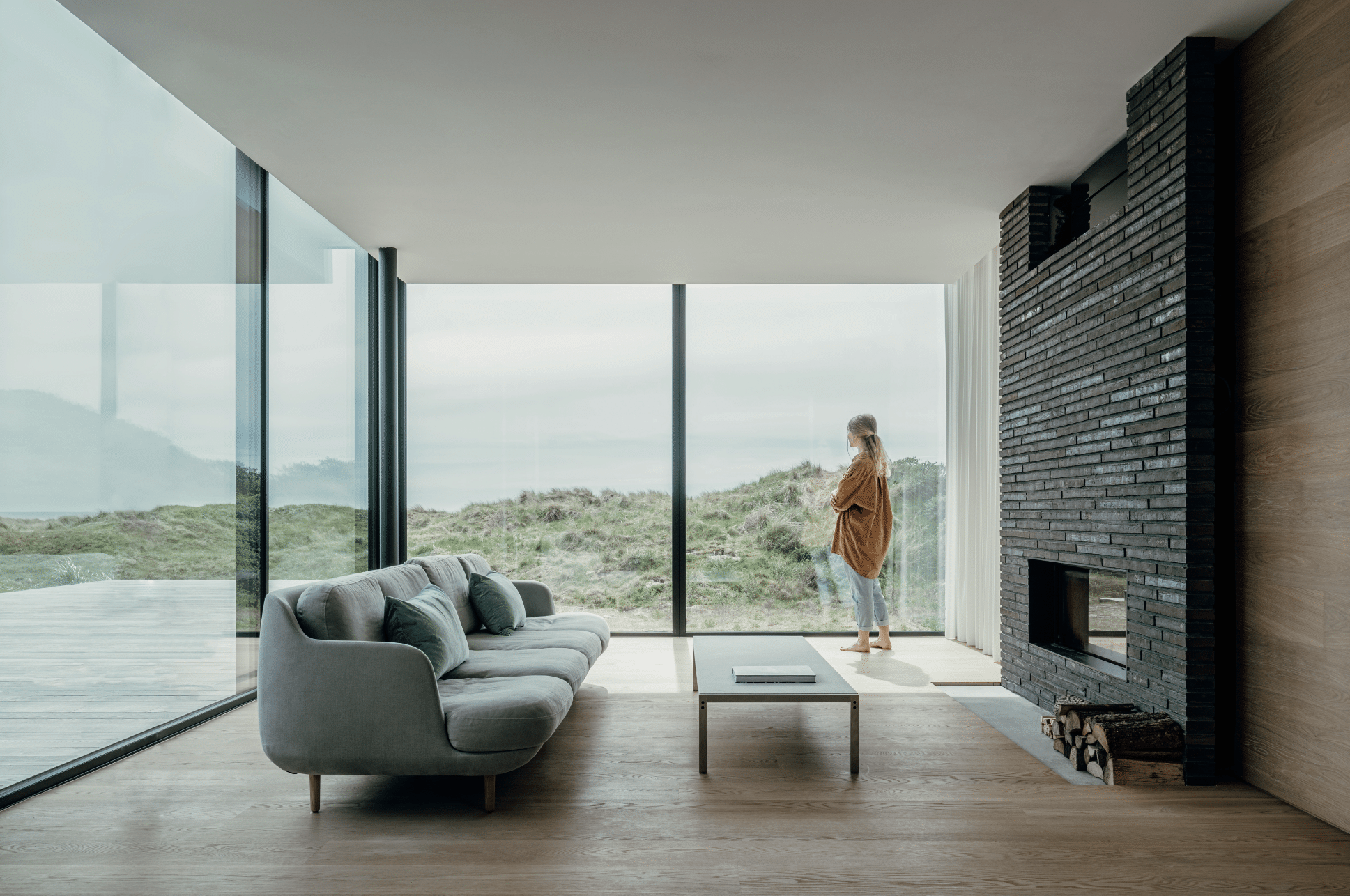 Solarlux zeigt ein Wohnzimmer mit Holzboden und einer großen, grauen Sitzlandschaft und einer dunklen Ziegelwand mit Kamin und einer rundherum Verglasung mit Aussicht ins Grüne.