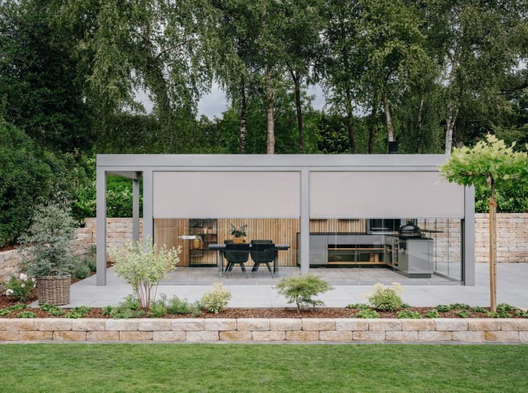 Solarlux zeigt einen Garten mit kleiner Ziegelmauer und überdachter Terrasse mit Markisen als Sonnenschutz und Verglasung, mit einer Outdoorküche und gemütlicher Sitzgruppe.