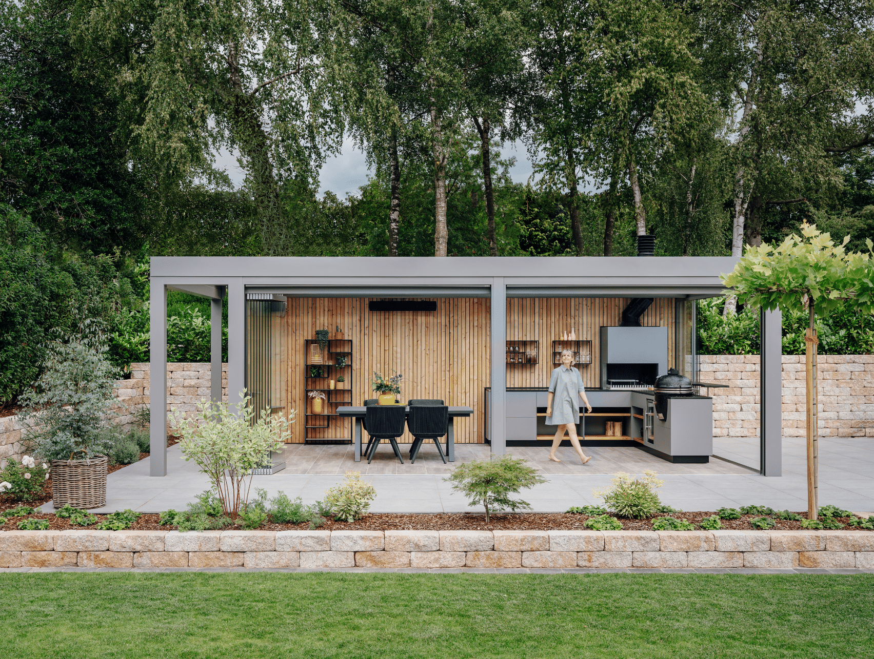 Solarlux zeigt einen Garten mit kleiner Ziegelmauer und überdachter Terrasse mit Holzverkleidung an der Rückwand und Verglasung, mit einer Outdoorküche und gemütlicher Sitzgruppe.