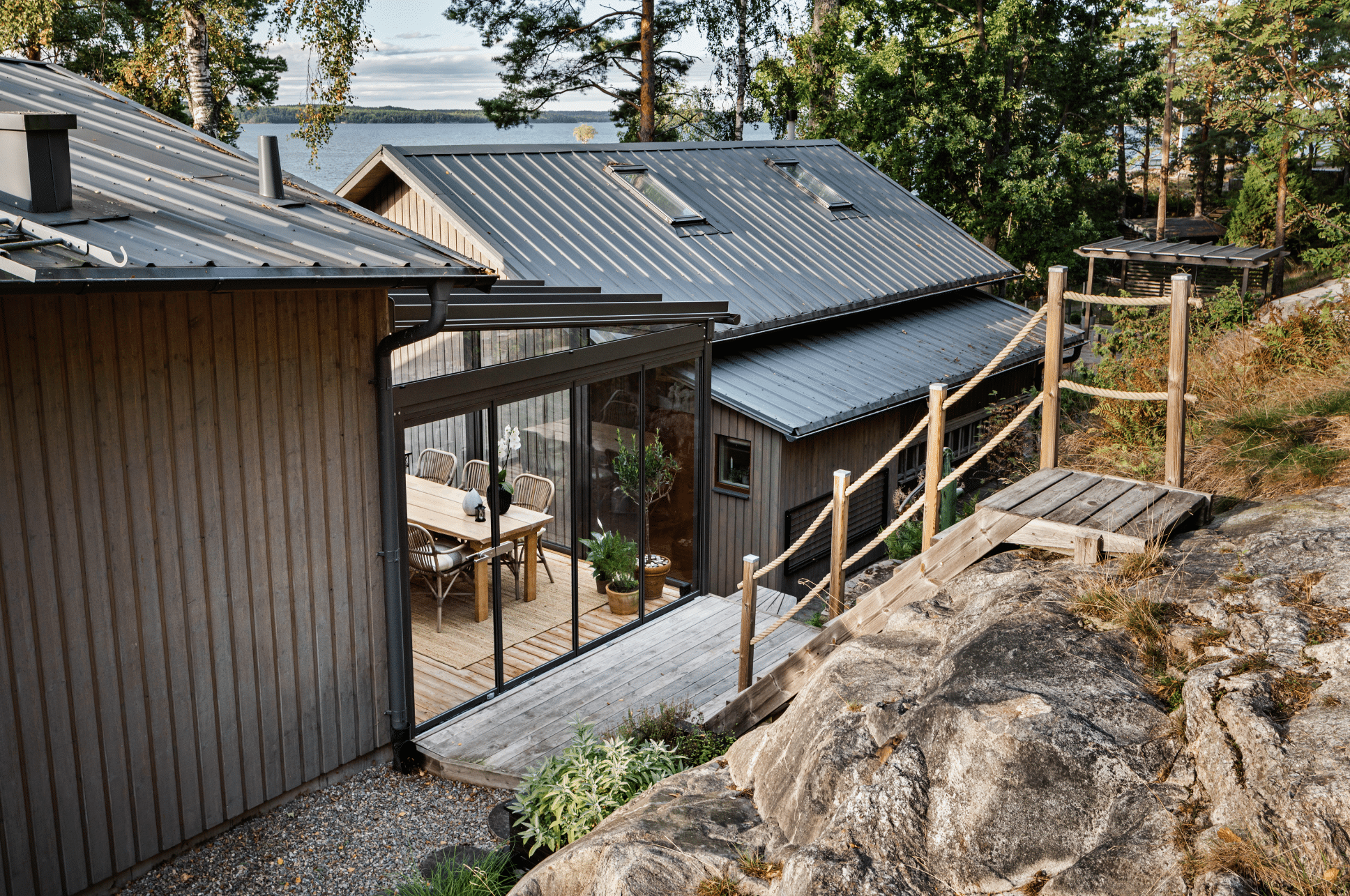 Solarlux zeigt ein Holzhaus mit großen, verglasten Wintergarten, einer Glasschiebetür mit Zugang  zu einer kleinen Terrasse aus Holz die zu einer schmalen Holztreppe führt.