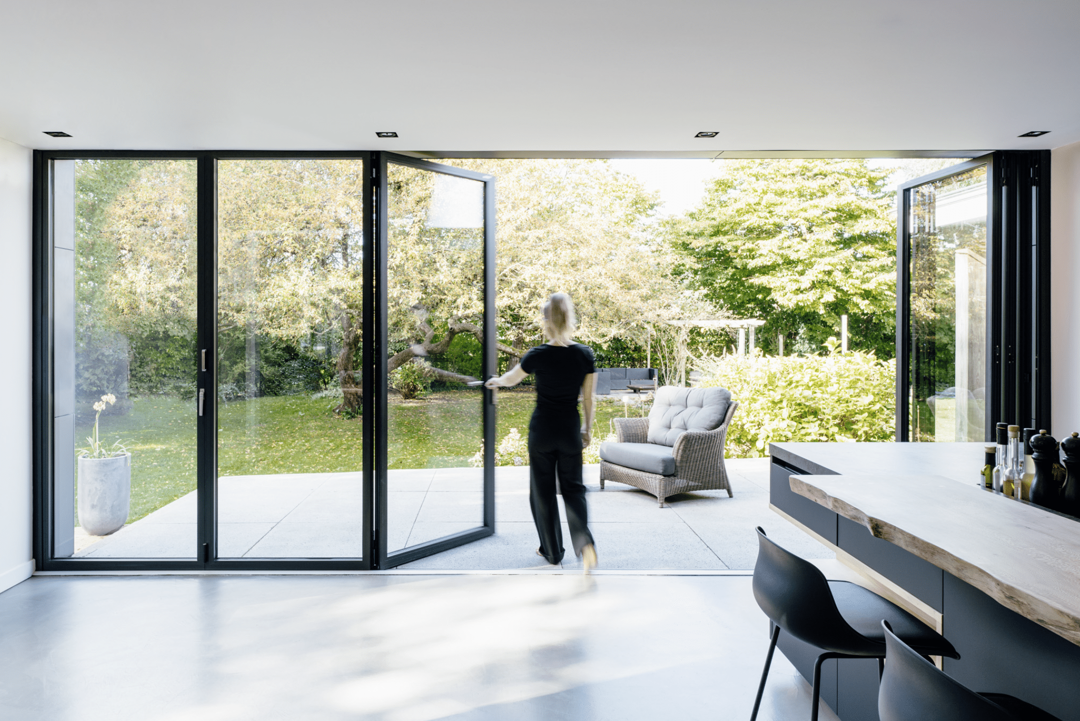 SOLARLUX präsentiert ein Glasfaltfenster, welches von der schwarzen Küche mit Holzelementen auf die Terrasse mit Outdoormöbeln führt.